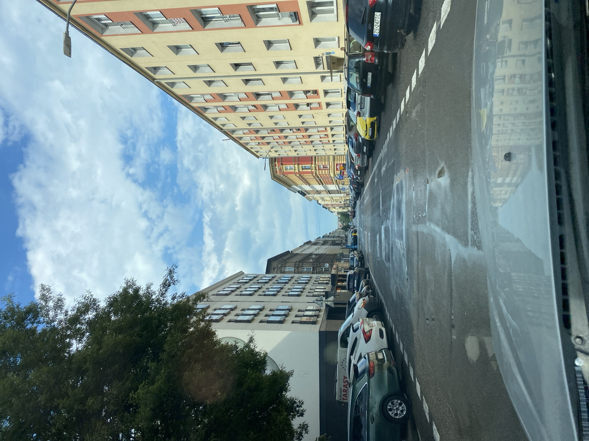 The image depicts a street scene in Szczecin, Poland. Tall, multi-story buildings with many windows line the left side of the street, some with cream-colored facades and others with reddish-brown accents. A lush green tree is visible in the foreground on the left. On the right side of the street, cars are parked along the curb. The road appears wet, suggesting recent rain or a damp surface.
Cars of various colors, including black, yellow, white, and dark green, are parked in a row. Some license plates are partially visible, one reading "ZS 247 TC". In the distance, more buildings and cars suggest a densely populated urban area. The sky is blue with scattered white clouds, indicating daytime. The perspective is from inside a vehicle, looking out at the street and buildings. Reflections on the surface of the vehicle on the right show buildings opposite. The overall atmosphere is that of a typical city street during the day, likely with moderate weather.
The text "TARASY" and "BUDIENIY" is visible on the side of a dark-colored van or truck parked on the right side of the street.