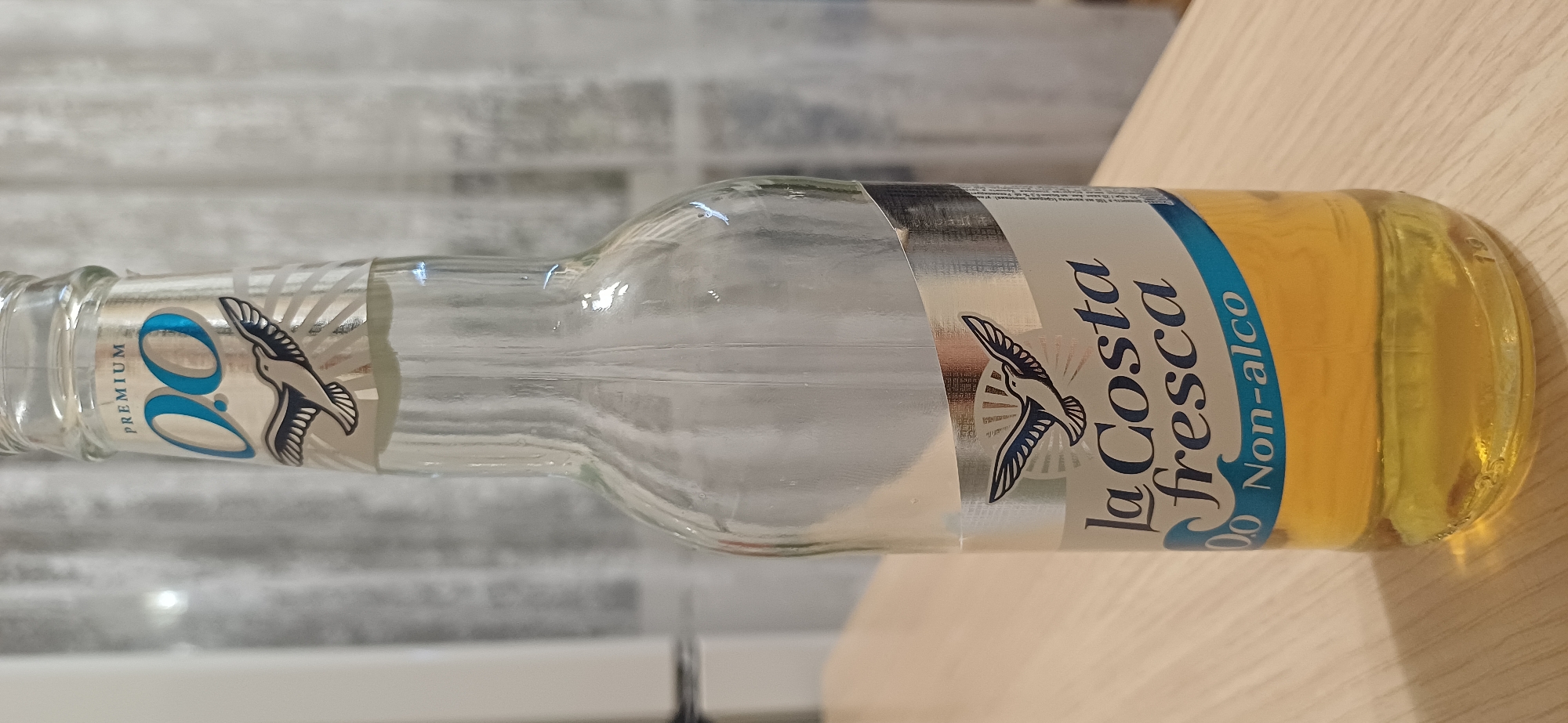 A clear glass bottle containing a light yellow liquid stands upright on a wooden surface. The bottle has a label with text and a graphic of a bird in flight. The background consists of sheer, light-colored curtains. The image is a close-up shot, focusing on the bottle. There is no information in the image to suggest it is located in Zuevka, Russia.