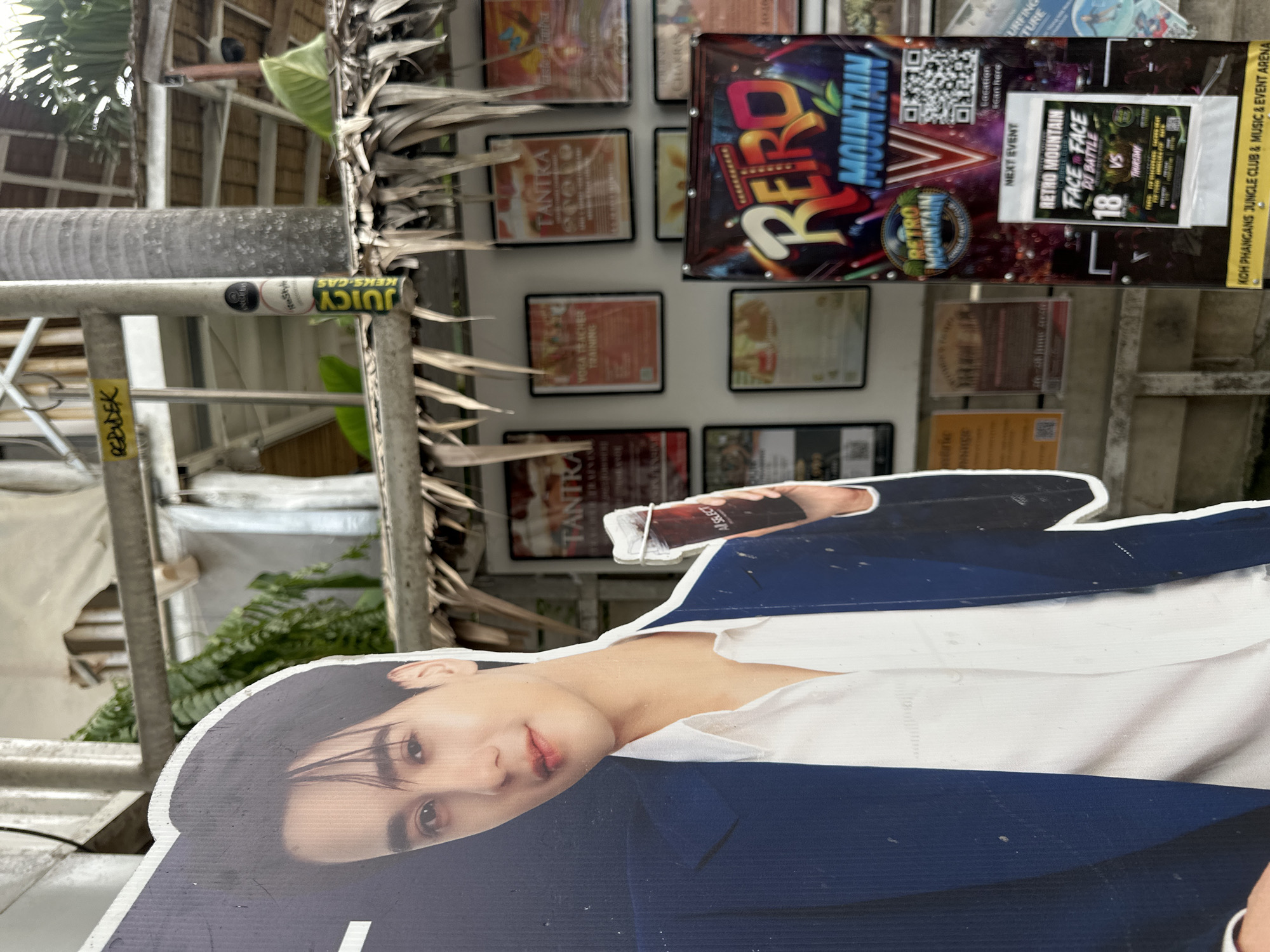 The image depicts an outdoor scene in front of a building, likely a venue, located in ตำบลเกาะพะงัน, Thailand. In the foreground, a large cardboard cutout of a young man wearing a dark blue suit and a white shirt is prominently displayed. He is holding a dark beverage in his right hand. To the right of the cutout, a large banner with the words "RETRO MOUNTAIN" in colorful lettering is visible. Below this banner, a smaller poster advertises an event titled "RETRO MOUNTAIN FACE 2 FACE DJ BATTLE" for the 18th of a month, which is a Thursday. Several framed posters advertising "TANTRA" and "YOGA TEACHER TRAINING" are mounted on the wall behind the cutout. The background shows tropical vegetation, including palm fronds, and parts of the building's structure, suggesting an open-air or semi-outdoor environment. A metal pole with various stickers is also present. The overall impression is that of a promotional display for an entertainment or wellness venue in a tropical setting.