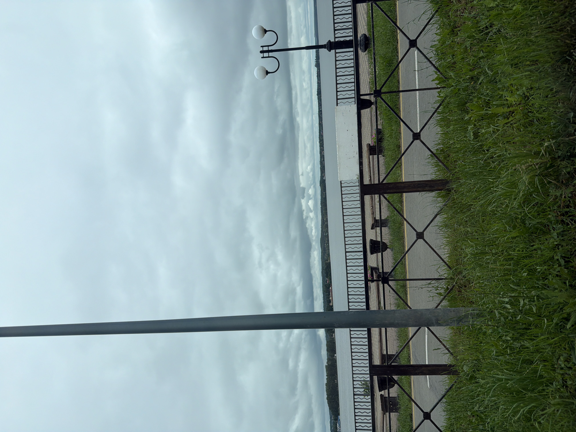 This image depicts a waterfront scene in Votkinsk, Russia, under an overcast sky. The upper half of the frame is dominated by a uniform expanse of grey, cloudy sky. A large body of water, likely the Votkinsk Pond, stretches horizontally across the middle ground, its surface appearing calm and grey. The far bank of the water features a horizon line of dense green trees, with hints of distant buildings or structures interspersed among the foliage.
In the foreground, a dark metal fence with a decorative wavy top rail and a criss-cross pattern in its lower sections runs horizontally, separating a grassy area from a paved path. Behind this fence, a paved promenade made of light-colored interlocking pavers is visible, dotted with several dark-colored benches or planters, and one distinct white rectangular block, possibly a bench or barrier. To the far right, a traditional black streetlamp with two spherical white globes extends upwards from behind the fence. A tall, slender, grey cylindrical pole occupies the left side of the frame, extending vertically from the bottom edge to the top edge, partially obscuring the view.
Below the metal fence and the paved promenade, an asphalt road or pathway is visible, marked by a thin white dashed line centrally and a thin yellow line to its right. The immediate foreground is covered with lush, green grass and various weeds. No individuals or animals are present in the scene. The overall lighting suggests daytime with heavily diffused light due to the dense cloud cover.
