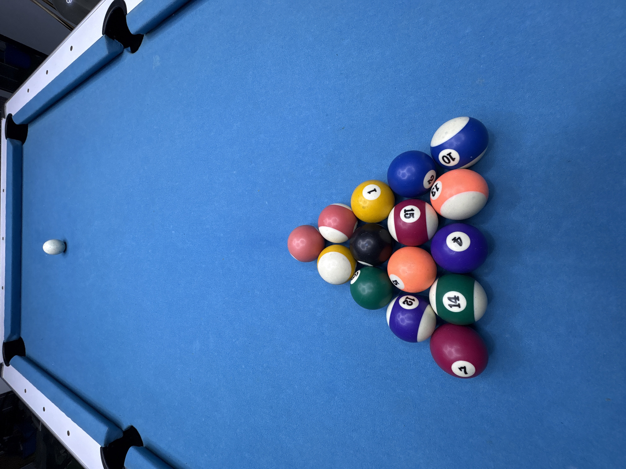 The image shows a top-down view of a pool table with a light blue felt surface. A set of fifteen pool balls is racked in a triangular formation on the right side of the table. A white cue ball is positioned on the left side, near the edge of the table. The table has white rails and black pockets. The location is unknown, and there is no evidence of Baan Bang Rak, Thailand.