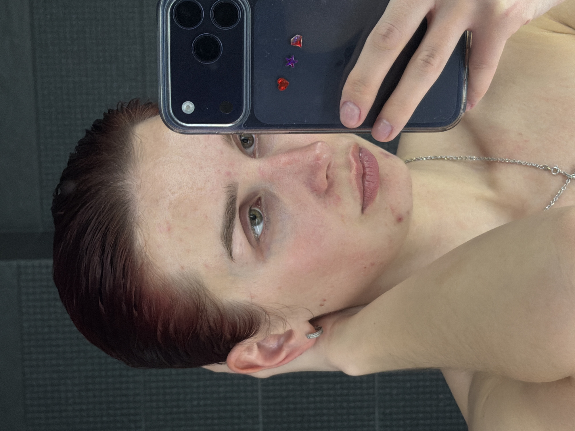 This mirror selfie, taken in Chaikovsky, Russia, features a person with reddish-brown, seemingly wet hair, fair skin, and green eyes. The individual's head is significantly tilted, appearing almost horizontal in the frame, as they look with a neutral expression towards the camera's reflection.
The person is wearing a thin silver chain necklace and a small silver hoop earring on their visible ear. They are holding a dark-colored smartphone, consistent with an iPhone due to its camera module design, encased in a clear protective cover. Adorning the back of the phone case are three small, shiny, colorful decorations: a red heart, a purple star, and an irregular red shape.
The background, visible in the reflection behind the person, consists of dark gray, rectangular tiled walls, suggesting an indoor setting such as a bathroom or a changing facility. Visible on the person's face are some areas of redness and small blemishes. The hair appears slicked back and wet, potentially indicating a recent shower or swim. No discernible text is present in the image.