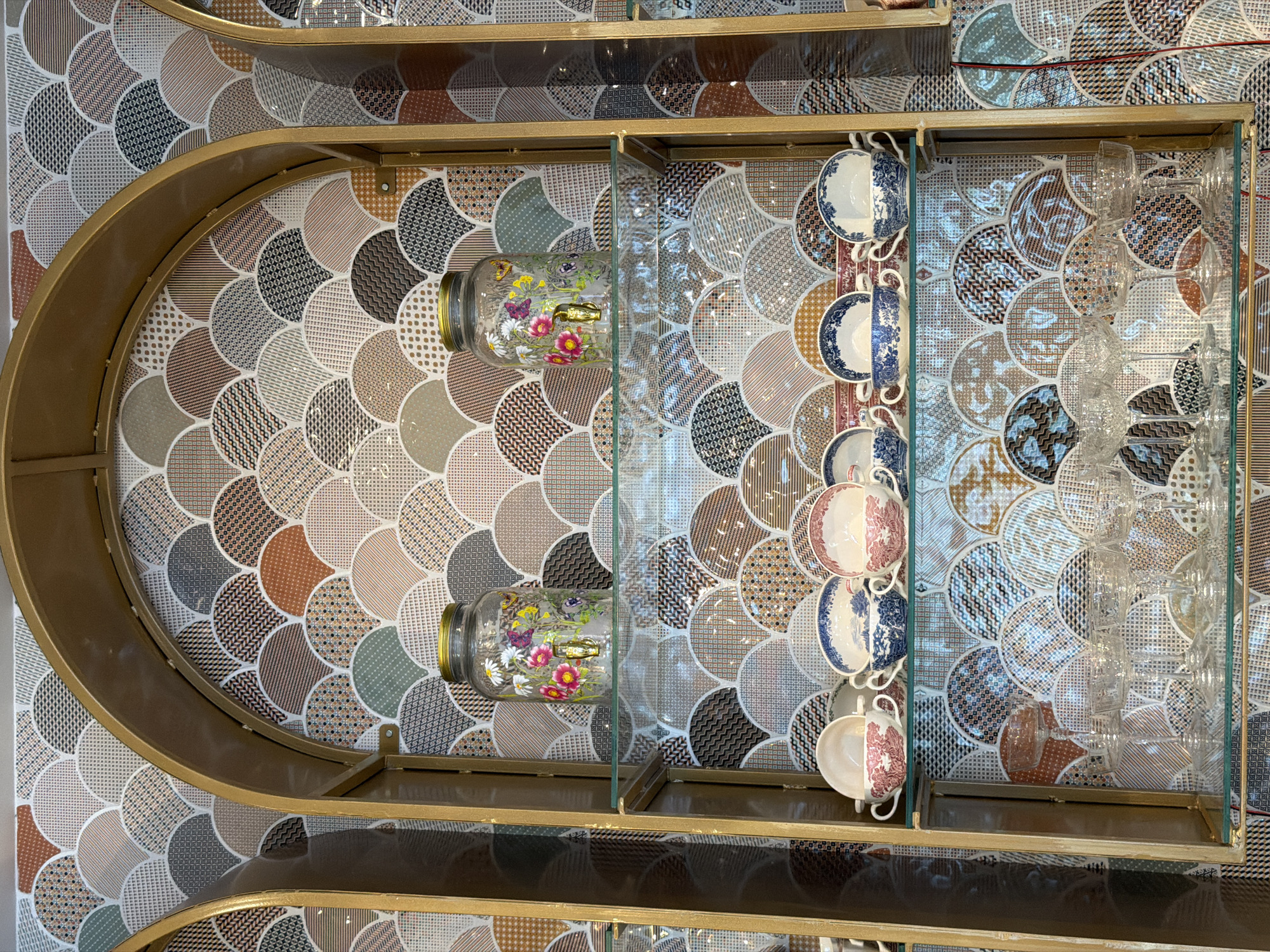 A high-angle shot depicts a shelving unit with a gold frame, separated by a glass panel, situated in Radovići, Montenegro. The shelf displays two glass jars filled with artificial flowers, arranged horizontally on the left side. To the right of the glass panel, a row of vintage teacups with a mix of blue, red, and white floral patterns is hanging on hooks. Further right, behind another glass panel, a collection of clear glasses, including what appear to be champagne flutes and drinking glasses, is arranged. The background behind the shelving is covered in a patterned mosaic tile design composed of semicircles with various textures and colors. The lighting is bright, highlighting the textures and colors of the mosaic tiles.