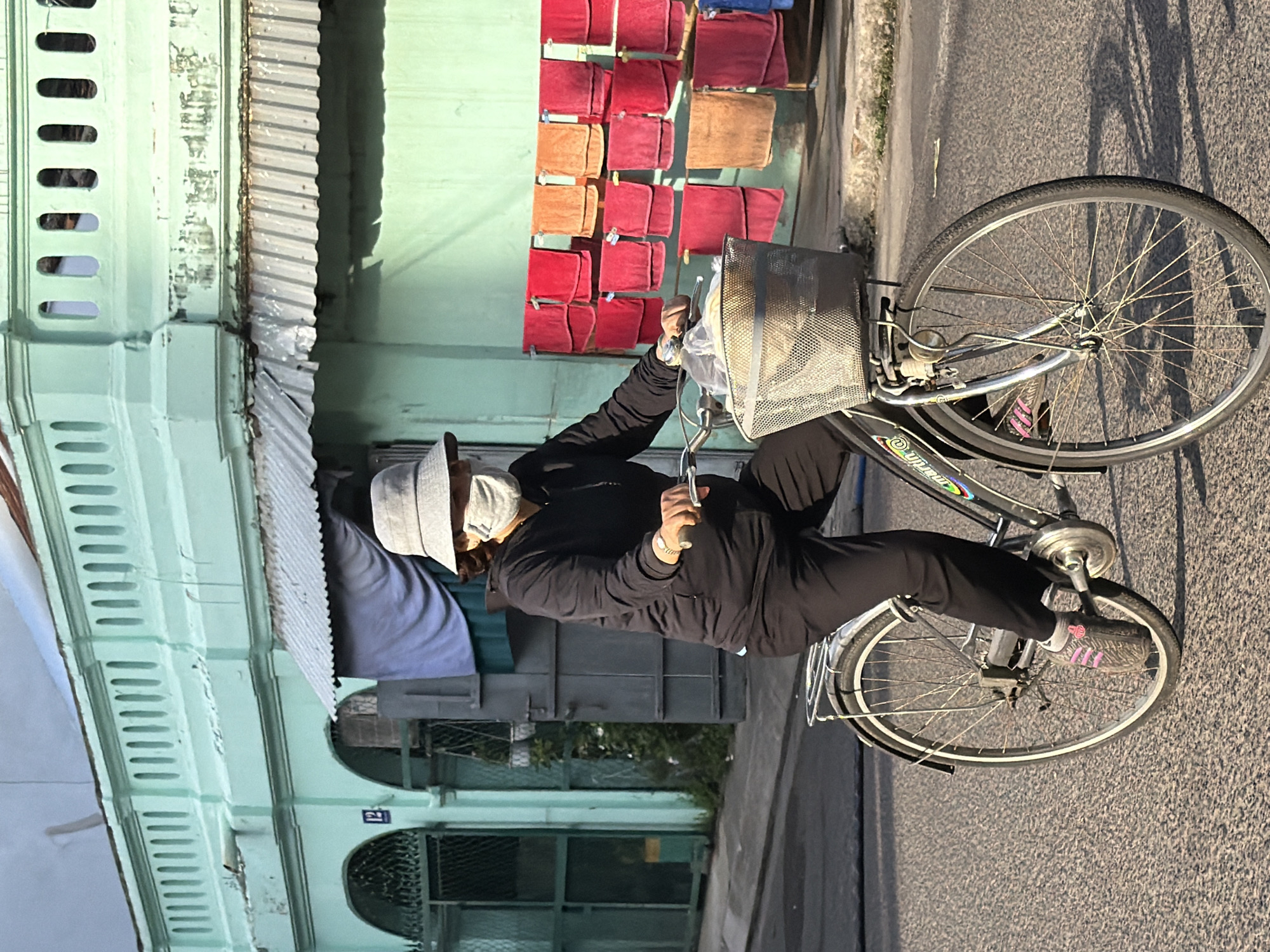 In Nha Trang, Vietnam, a woman wearing a grey bucket hat and a face mask rides a bicycle down a paved street. She is dressed in a black hooded sweatshirt and black pants. The bicycle has a metal basket on the front, filled with plastic bags. In the background, a light mint green building with decorative architectural details stands, with several rows of red and orange towels hanging on a line outside. The scene is illuminated by natural sunlight, casting shadows on the road.