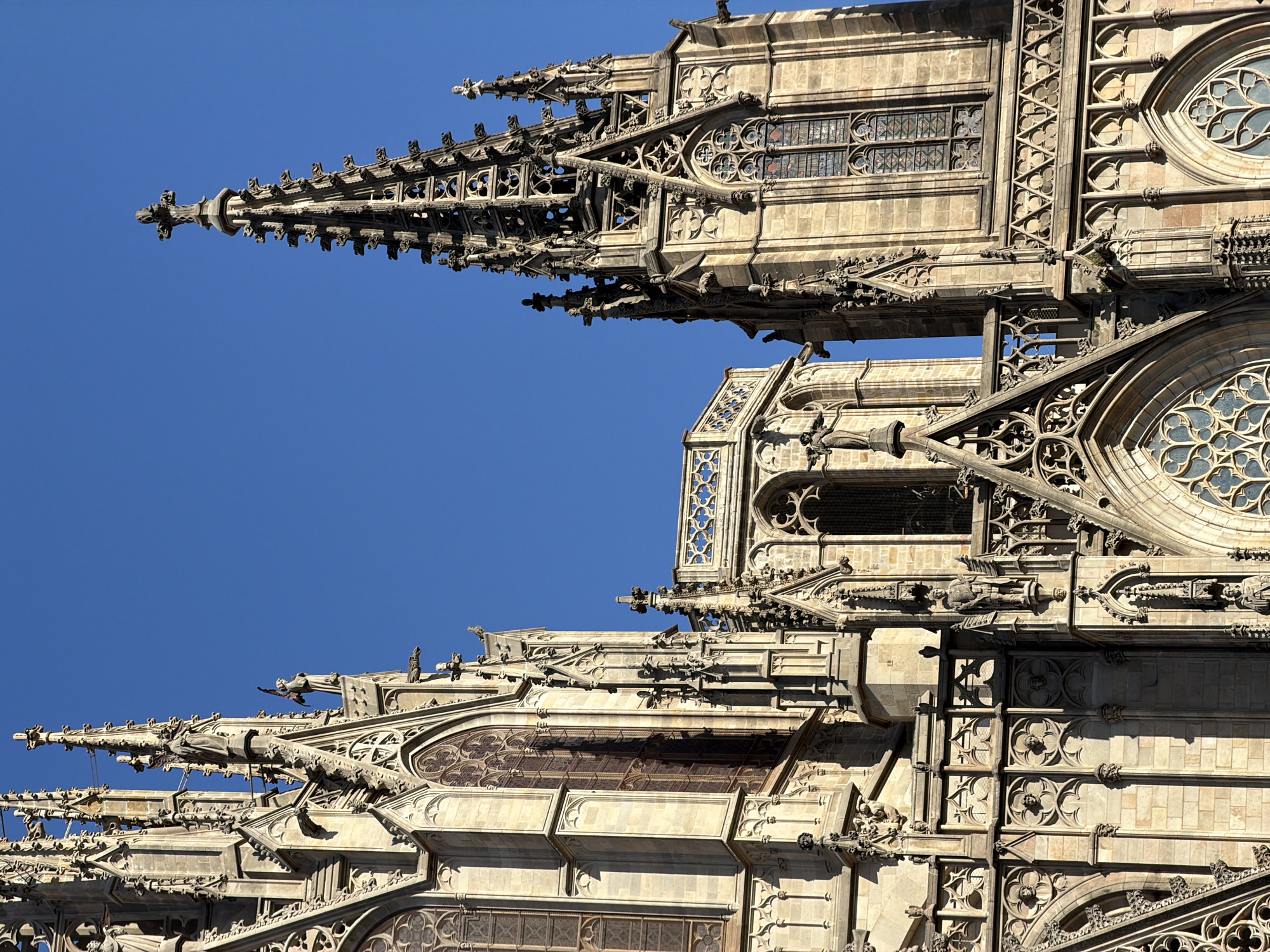 The image shows the facade of the Barcelona Cathedral, a Gothic cathedral located in the Barri Gòtic (Gothic Quarter) of the city of Barcelona, Spain. The photograph captures a close-up view of the intricate architectural details of the cathedral's towers and spires against a clear blue sky. The stonework is elaborate, featuring pointed arches, stained-glass windows, gargoyles, and sculptures of religious figures. Sunlight casts distinct shadows, highlighting the three-dimensional aspects of the facade. The composition focuses on the upper portions of the cathedral, emphasizing its height and the craftsmanship of its construction. There are no people visible in the image.