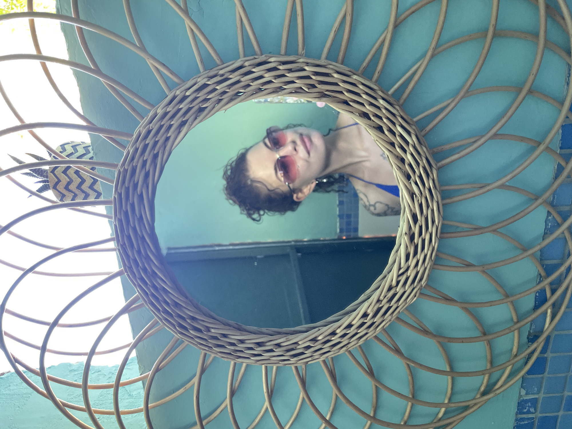 A woman with curly brown hair pulled up in a bun and pink sunglasses is reflected in a round, woven rattan mirror. She is wearing a blue bikini top. She has a tattoo on her left arm. The mirror is framed by a large, decorative sunburst pattern made of woven rattan. The background is a teal-colored wall with blue mosaic tiles at the bottom. A decorative object resembling a pineapple with a chevron pattern is visible in the upper right of the reflection. The scene appears to be an outdoor setting, possibly a patio or garden, given the bright sunlight. The location is identified as Prijevor, Montenegro.