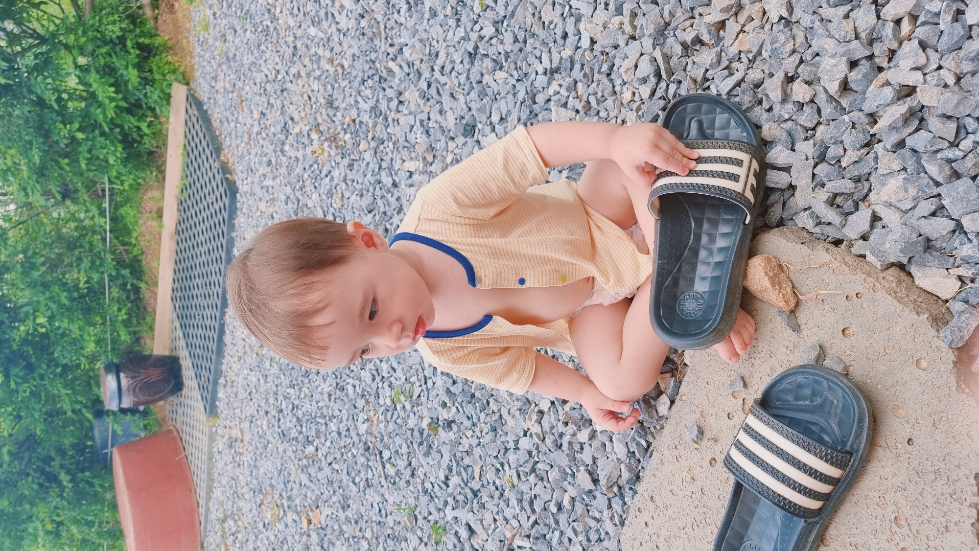 A young child sits on a gravel surface with two sandals near the child's feet. The child is wearing a light yellow shirt with dark blue trim. The gravel area is next to a patch of green foliage, a grated surface, and a red bucket. This photo was taken in Dunnae-myeon, South Korea.