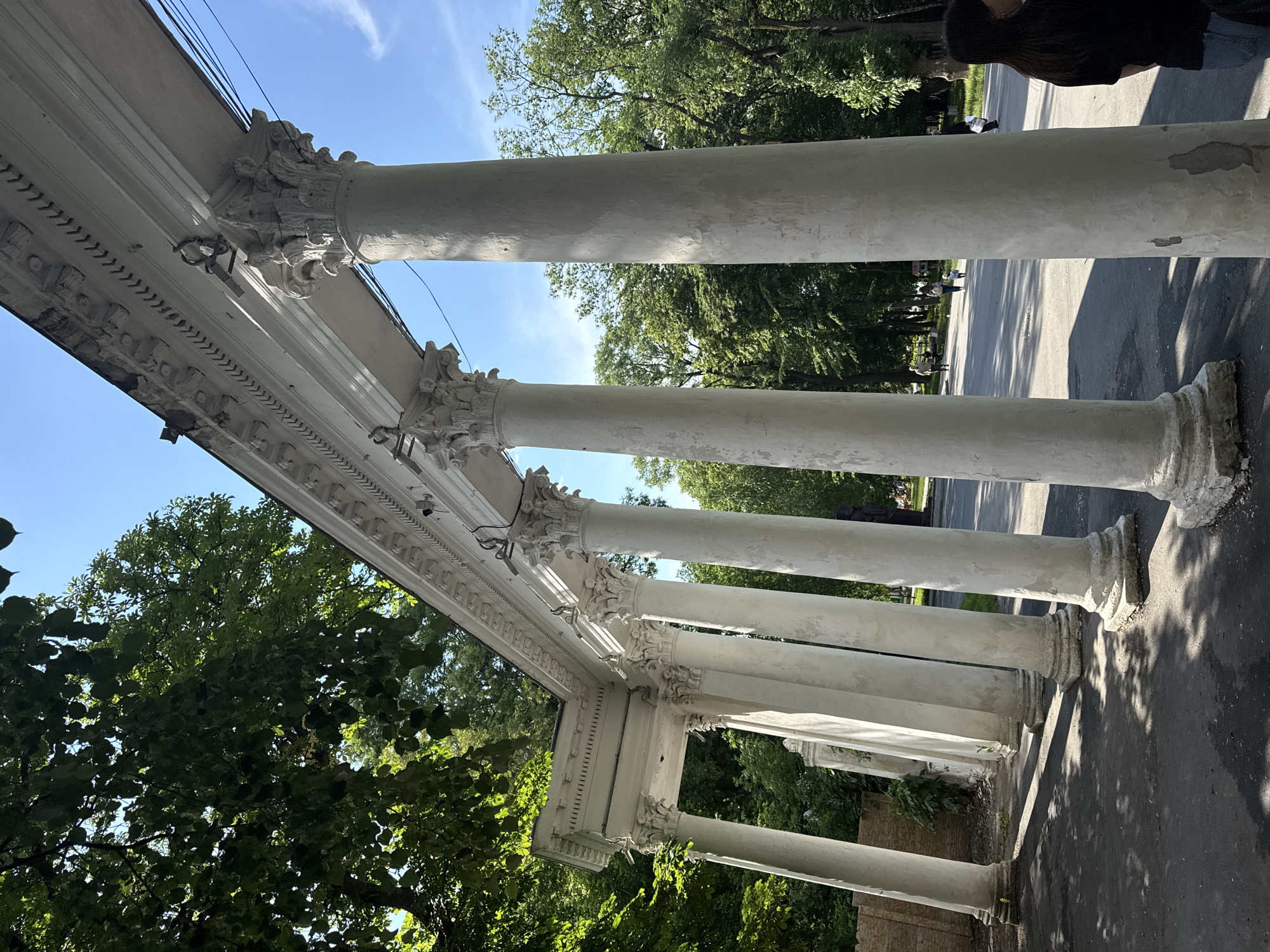 The image depicts an outdoor scene featuring a long, white colonnade with Corinthian-style columns, suggesting an architectural element within a park or public space. The colonnade is viewed from a low angle, emphasizing its height and the intricate details of its capitals and entablature. Sunlight casts dappled shadows on the asphalt path in the foreground, indicating a bright, possibly sunny day. Lush green trees with dense foliage flank the colonnade and extend into the background, creating a naturalistic setting. Several indistinct figures are visible in the middle ground, walking along a path beyond the colonnade. The sky is a clear, vibrant blue with a few wispy clouds. The architecture and the surrounding greenery suggest a location such as the National Pirogov Estate Museum in Vinnytsia, Ukraine, which features similar classical architectural elements and parkland.