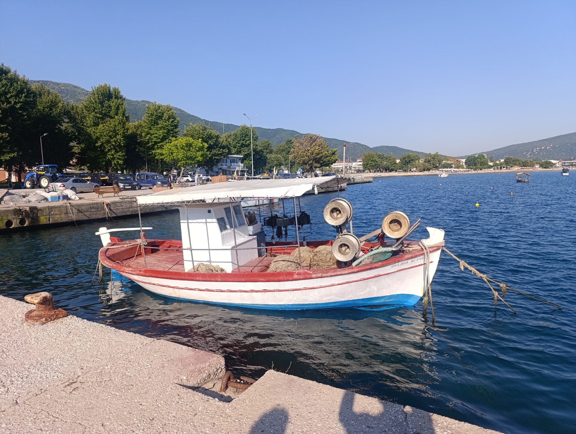 In Σταυρός, Greece, on a clear, sunny day, a small white fishing boat with a red and blue hull is moored to a concrete pier. The boat has a white cabin and a canopy. Fishing nets and coiled ropes are visible on deck, along with two large fishing reels mounted at the stern. The water is a deep blue and slightly rippled. In the background, a treelined shore with buildings and mountains can be seen. Cars and a blue tractor are parked along the waterfront, and a few other boats are visible in the distance. The scene is calm and depicts a typical harbor setting.