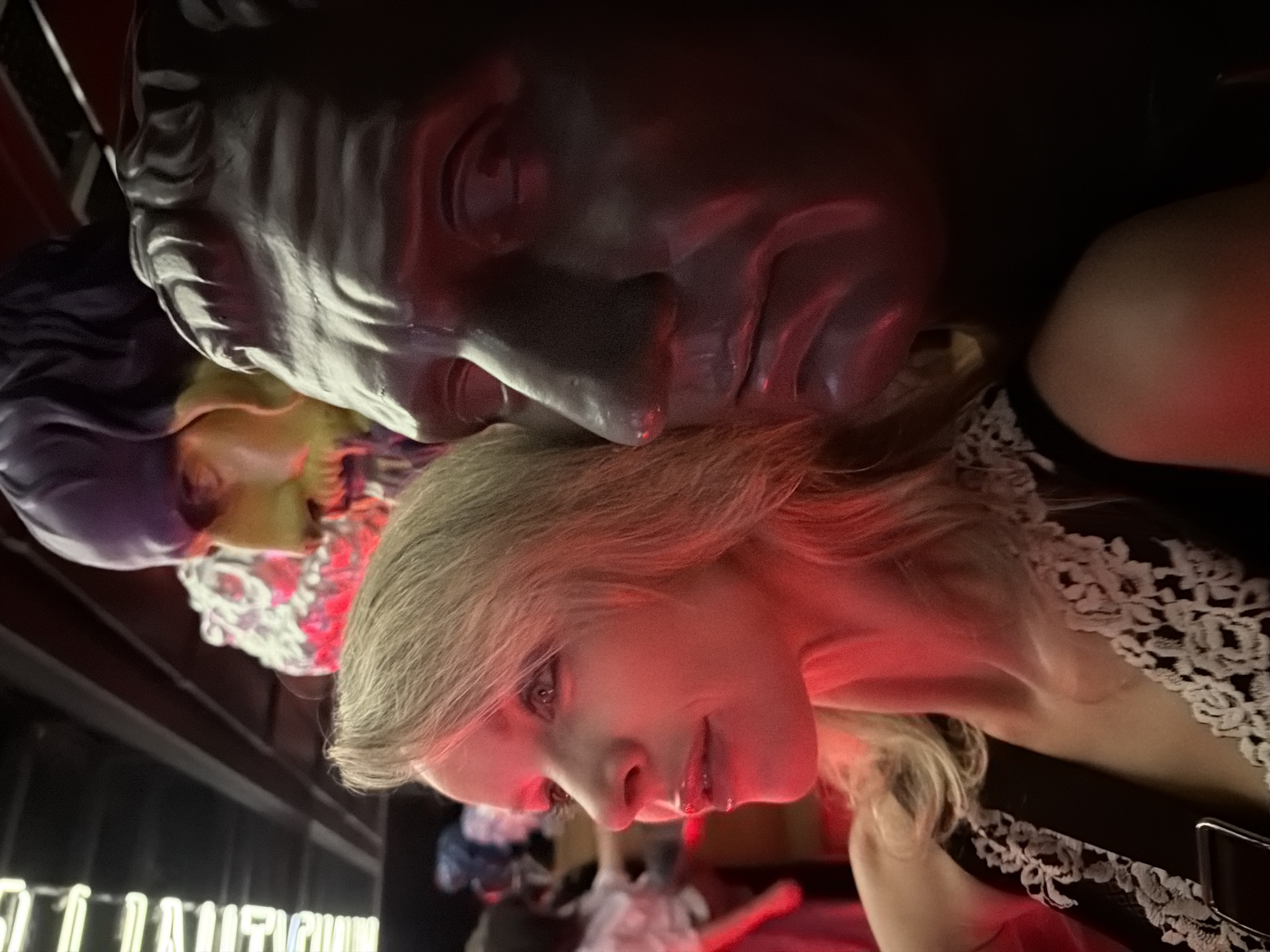 The image shows a woman with blond hair and pale skin leaning her face against a large, gray-colored bust of a man with a classical or Roman appearance. The scene is illuminated with red light. A second, smaller bust, colored in purple, yellow, and white, is visible behind the blond woman. The woman is wearing a black dress with a white lace trim. In the background, there are blurred figures and illuminated lettering that partially spells out "CAUTION." Given the style and general appearance, it is possible that this image was taken in a stylized or artistic environment. Without further information, it is not possible to confirm that the image was taken in Moscow, Russia.