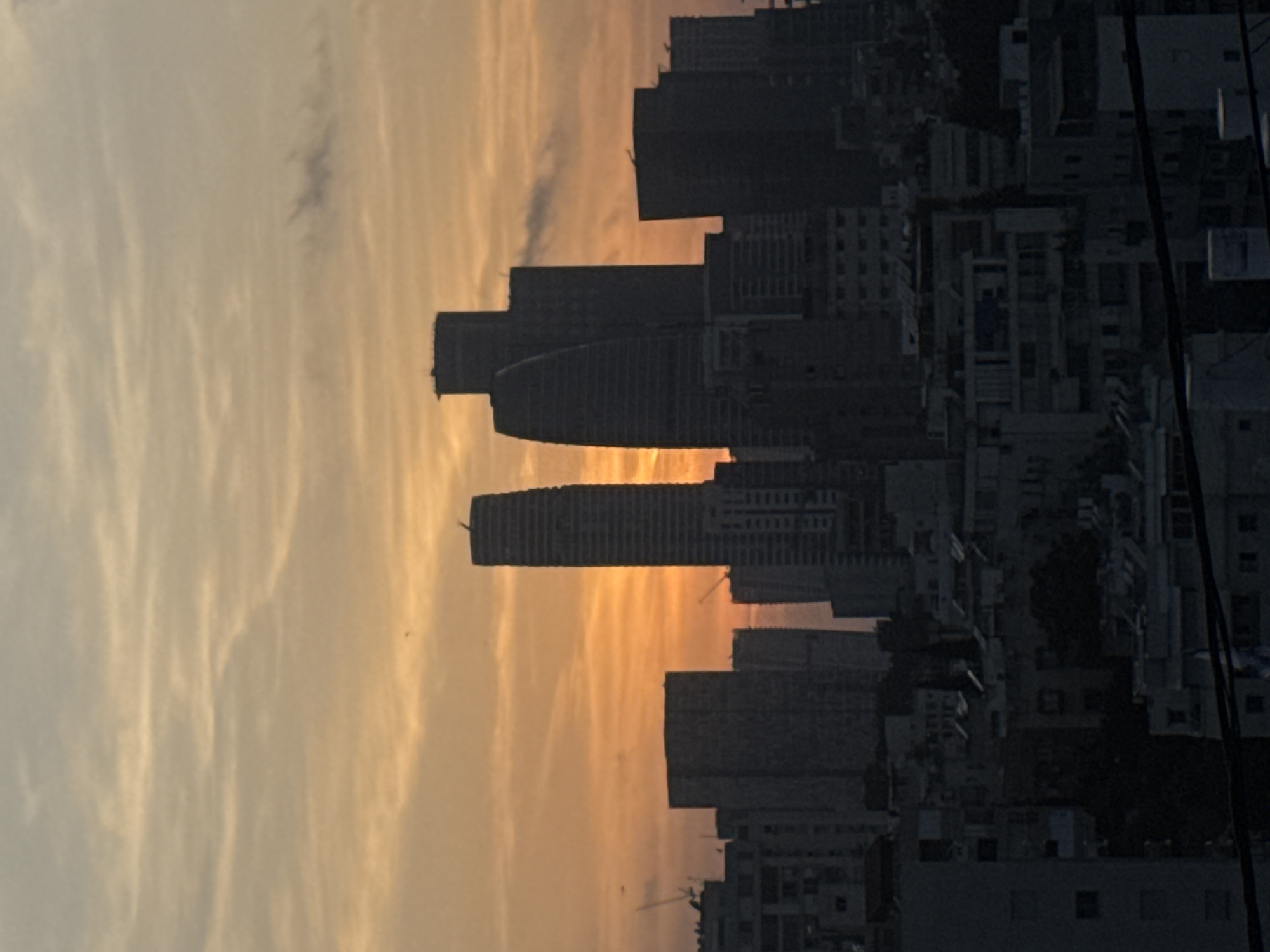 The image depicts the Ramat Gan skyline in Israel at sunset. Tall buildings of various heights are silhouetted against an orange and yellow sky. In the foreground are residential buildings, creating a sharp contrast between the buildings and the sky. Electrical wires are partially visible on the right side of the image.