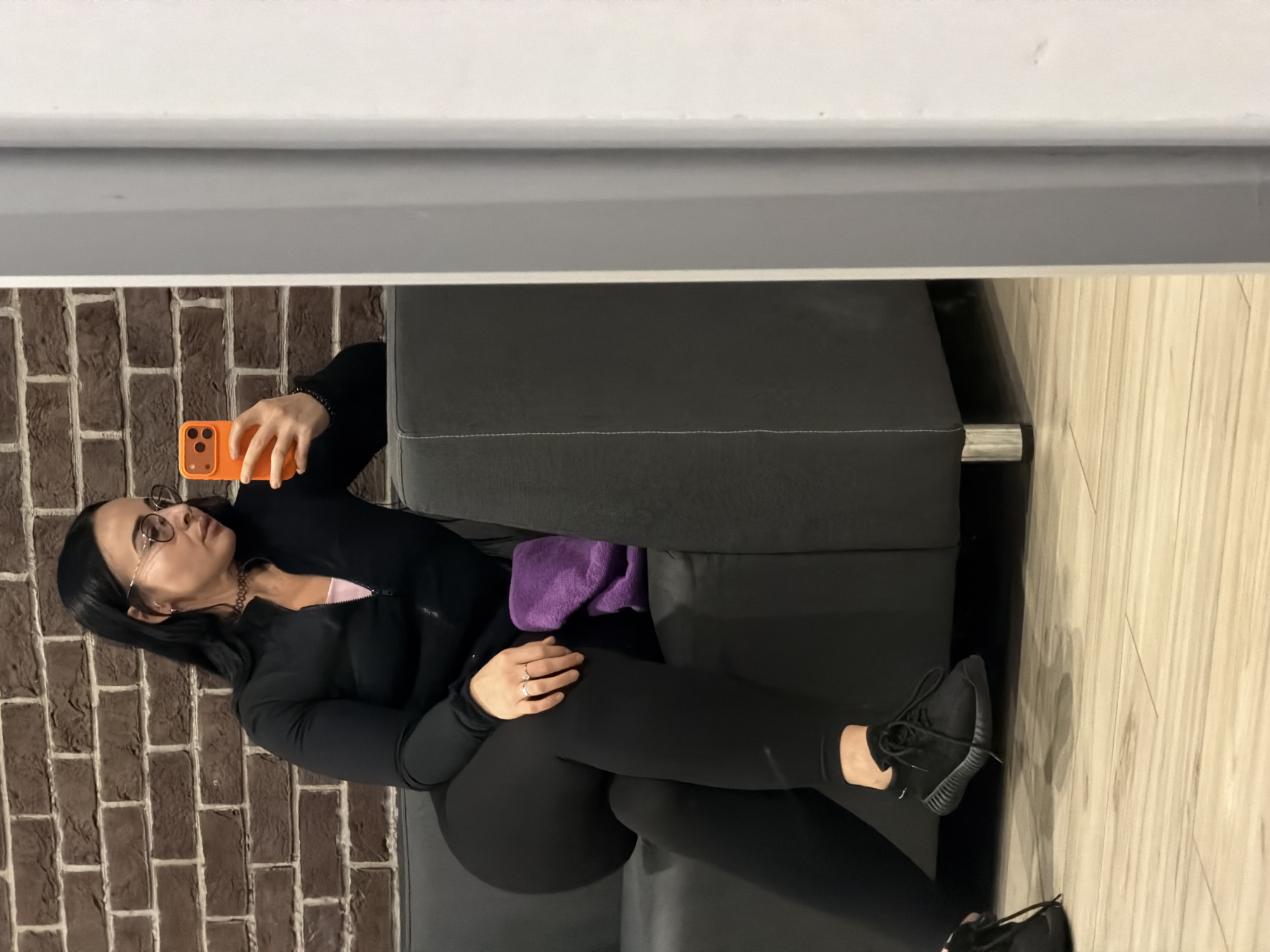 The image displays a woman relaxing indoors, reclining on a dark grey or black couch. She has dark hair and is wearing glasses, looking upwards and slightly to her right. Her attire includes a black long-sleeved zipped top, possibly over a pink inner top, black leggings, and black athletic shoes. A black choker necklace is visible around her neck, and she wears a ring on her left ring finger.
She is propped up on her right elbow, with her left leg bent and her right leg extended. In her left hand, she holds an orange smartphone, which features a triple-lens camera on its back. A purple towel or cloth is tucked beside her on the couch cushion. One black athletic shoe is on her left foot, while another shoe lies detached on the light-colored wooden or laminate flooring beside her feet.
The background behind the couch features a reddish-brown brick wall with light-colored mortar on the left side of the frame. Above the couch and to the right, a light grey or white wall or panel is visible. The indoor setting appears to be a lounge or waiting area, possibly in a gym or a public space within Saint Petersburg, Russia, given the contextual information. The lighting is ambient, suggesting an indoor environment without specific time of day indications, and there is no visible text. The woman appears relaxed, engrossed with her phone.