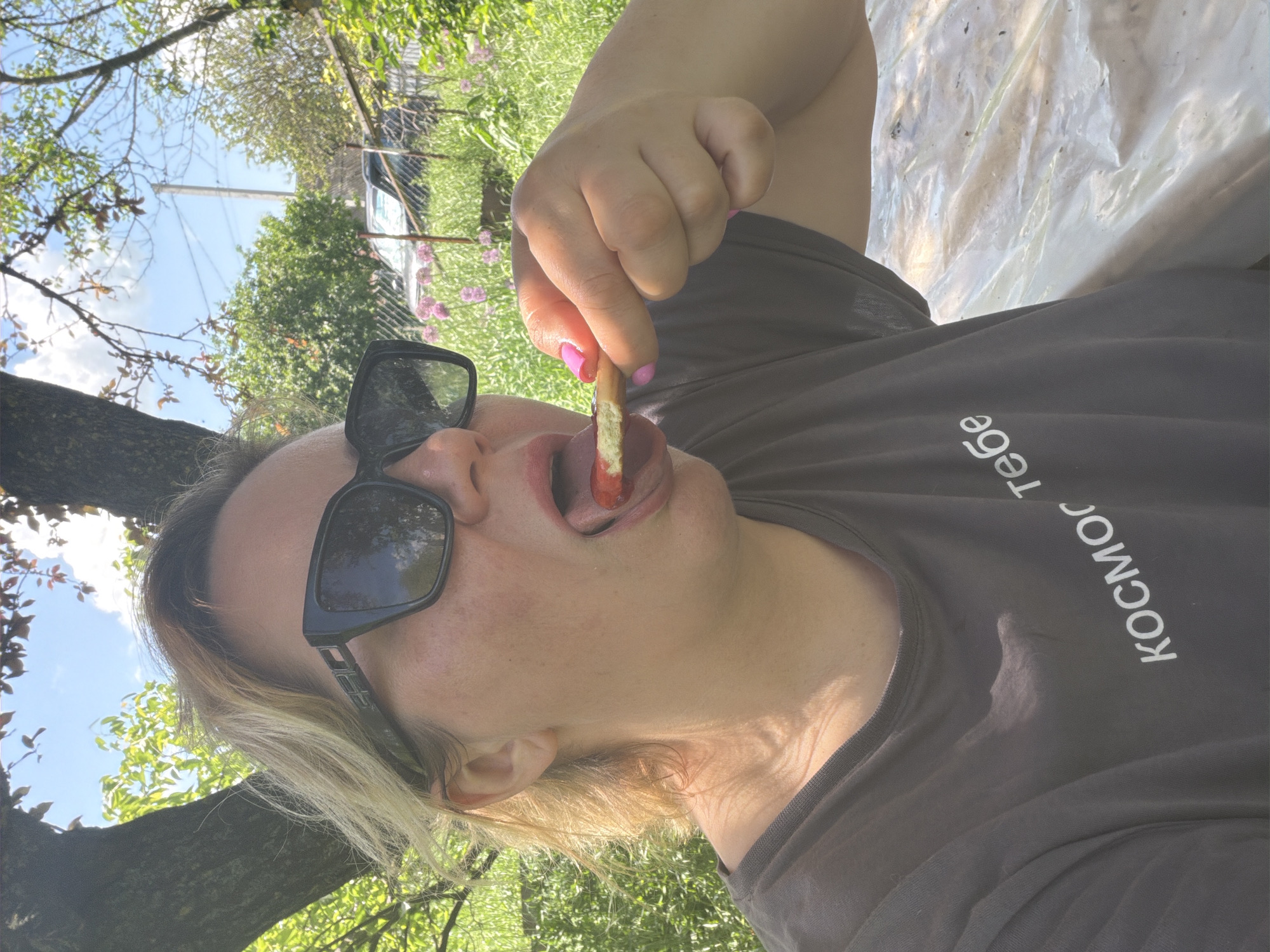 A woman is outdoors, possibly in Leningradskaya, Russia, holding a snack to her mouth. The woman is wearing dark sunglasses and a dark t-shirt with white Cyrillic lettering, the shirt seems to have sweat stains on it. She has pink nail polish. The background consists of trees and vegetation. A clear plastic sheet is also visible.