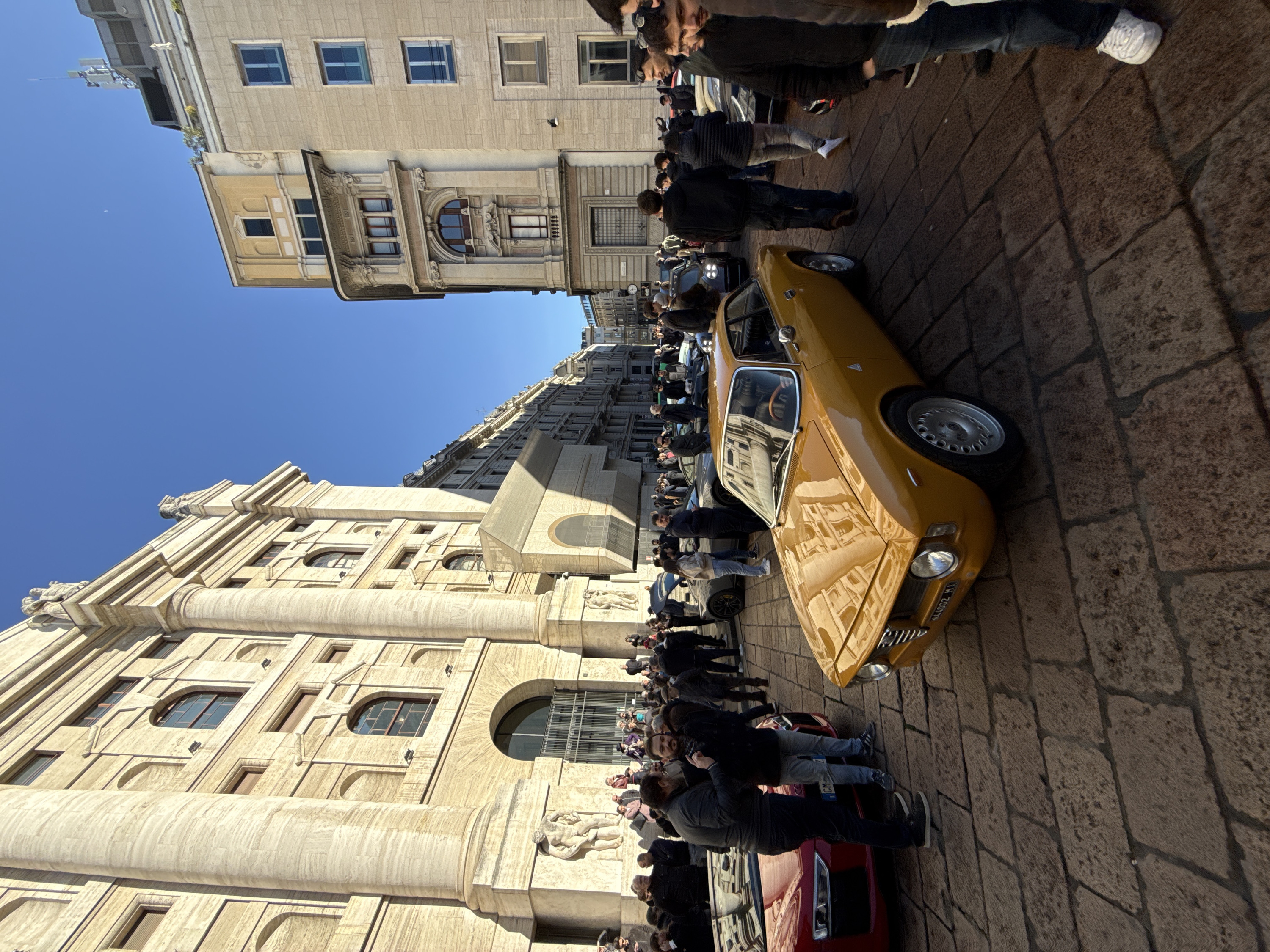 The image depicts an outdoor gathering of people and vehicles in Piazza Affari, Milan, Italy. The prominent building on the left is the Palazzo della Borsa, characterized by its large light-colored stone facade, colossal fluted columns, arched windows, and a sculpture of a male figure embracing a bull at its base.
In the foreground, a bright yellow (mustard-colored) classic Alfa Romeo sports coupe is parked diagonally, displaying its front grille, circular headlights, and multi-spoke silver wheels. To its left, a partially visible red Audi R8 sports car is parked. The ground is paved with large, irregular cobblestones or sett stones.
Numerous individuals are present, observing the vehicles and engaging in conversations. Many are dressed in casual outerwear such as jackets and sweaters. Two men are conversing near the red Audi R8, one in a dark jacket and jeans, the other in a dark jacket and light blue jeans. A dark-haired man in a black jacket is visible in the far right foreground, partially obscured. Various other sports cars, both classic and modern, are distributed throughout the midground, surrounded by groups of onlookers.
The right side of the scene features multi-story city buildings with various window styles, including arched and rectangular shapes. One building on the far right has a cellular antenna array mounted on its rooftop. The sky above is clear and blue, with a faint crescent moon visible in the upper right quadrant. The scene is illuminated by direct sunlight, casting strong shadows on the cobblestones and highlighting the architectural details of the Palazzo della Borsa.