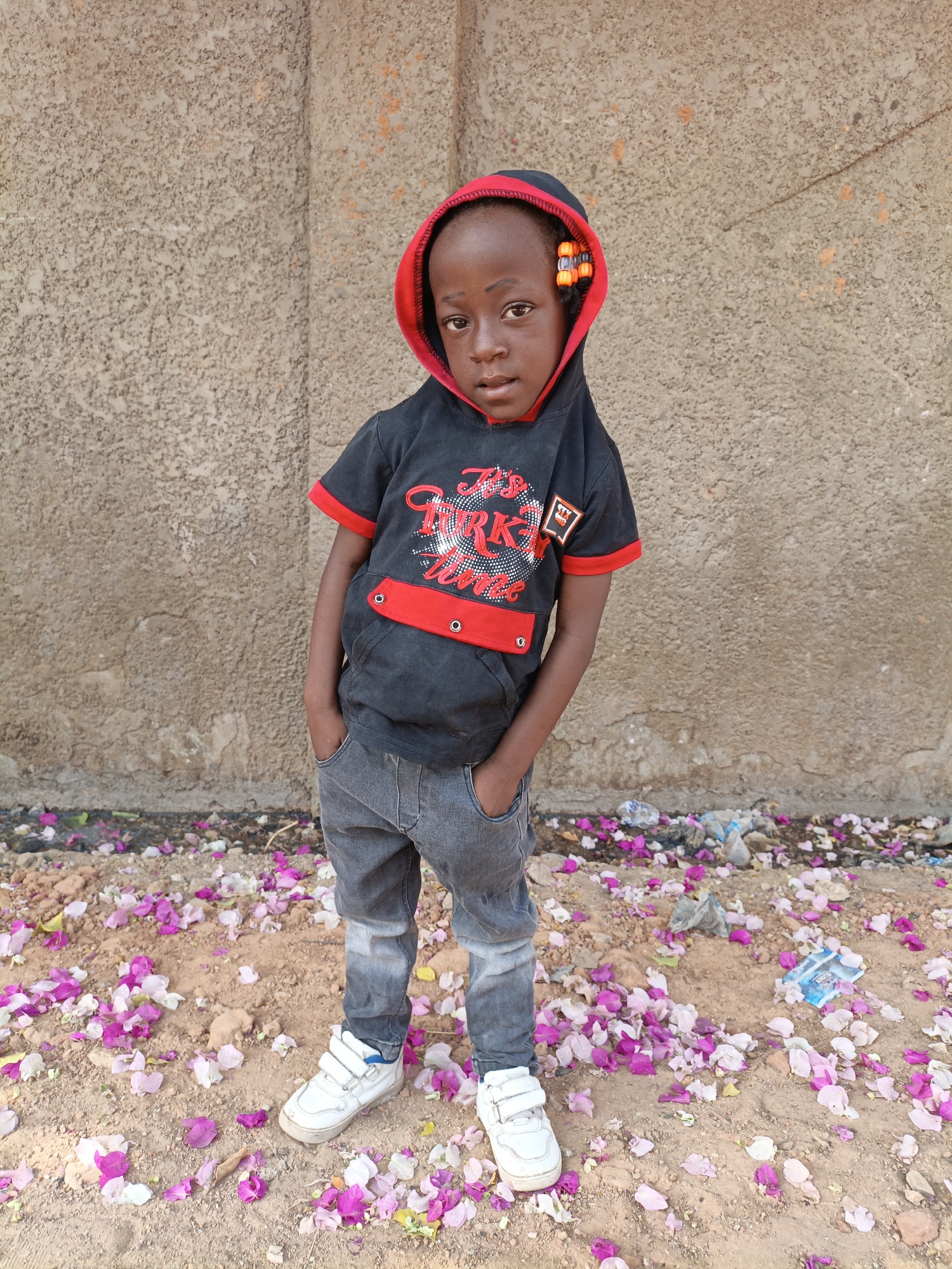 A child stands facing the camera in Gwagwarwa, Nigeria. The child wears a black hooded shirt with red trim and a graphic on the front, gray jeans, and white sneakers. The child has dark skin and wears beads in their hair. The child's hands are in their pockets. The ground is covered in loose soil and scattered pink and white flower petals. There is also trash on the ground. Behind the child is a textured, light brown wall.