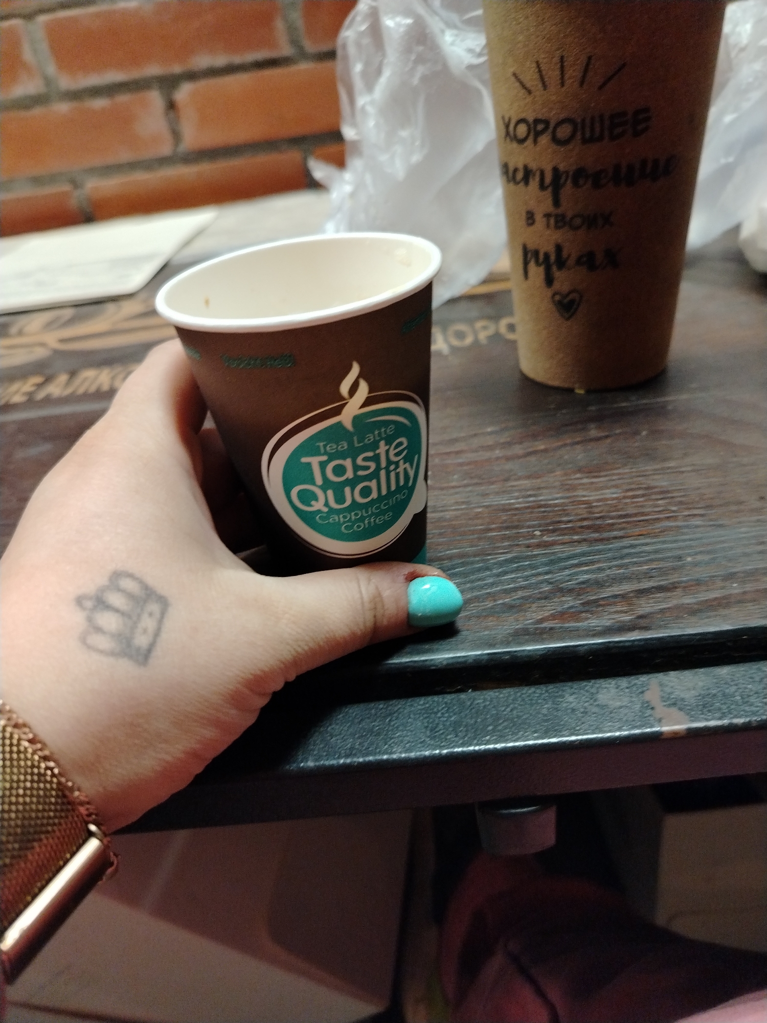 A close-up shot captures a person's hand holding a disposable coffee cup, placed on a dark wooden table or counter. The hand, with light skin, displays a small, faded crown tattoo on the back and a bright turquoise-painted thumbnail. A rose gold-colored mesh watch strap is visible on the wrist.
The brown paper cup being held has a white rim and a green and white logo that reads "Tea Latte Taste Quality Cappuccino Coffee" in English. In the background, another taller, craft paper-brown disposable cup stands, decorated with handwritten black text in Cyrillic. This text translates to "Good mood in your hands," reading "ХОРОШЕЕ настроение в твоих руках" (KHOROSHEYE nastroyeniye v tvoikh rukakh), and is accompanied by a small heart symbol.
A translucent plastic bag is partially visible behind the cups. The upper left background reveals a reddish-brown brick wall, suggesting an indoor setting such as a cafe, office, or home in Rusanovo, Russia. The scene depicts a casual moment, likely a coffee break, with a focus on the beverages. The lighting is even, without strong shadows, indicating artificial light or diffuse daylight.