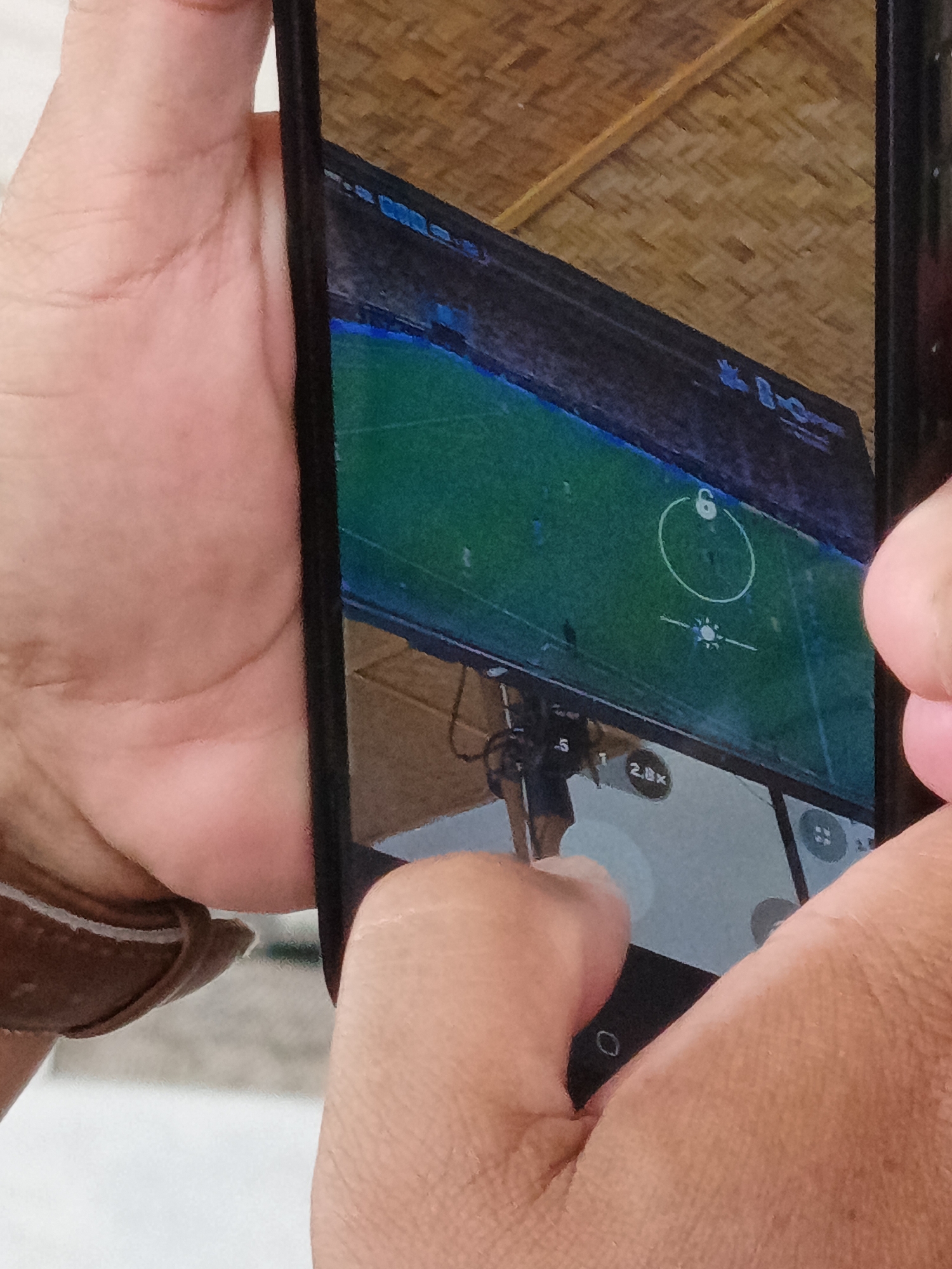 An overhead, close-up, vertical shot captures a person's hands holding a smartphone. The phone is oriented vertically, and its screen displays a live camera feed. The phone's screen is divided into two sections, with the top section showing a drone's perspective of a green field, possibly a soccer field, with blue markings and a yellow object in the center. A white circle with a "6" inside is visible on the field. The bottom section of the screen appears to be a camera interface, showing zoom options like "2.8x" and other icons. The background of the image consists of a woven bamboo ceiling and the hands of the person holding the phone, suggesting an indoor environment. The specific location in Julikeude Dua, Indonesia, is not discernible from the image itself.