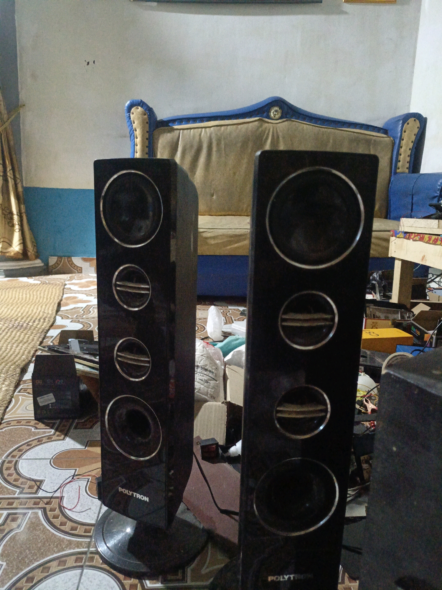The image shows two tall, black speakers on a patterned tile floor. The speakers have multiple circular drivers, some with metallic trim. The left speaker has "POLYTRON" written on it. Behind the speakers is a couch with blue and gold upholstery and a light-colored cushion. To the left of the couch, a curtain is visible. Various objects, including cardboard boxes and what appear to be electronic components, are scattered around the floor and on a low table behind the speakers. The wall behind the couch is light-colored with some dark marks. The scene appears to be an indoor residential setting, possibly a living room or entertainment area, in Pagar Ayu, Indonesia.