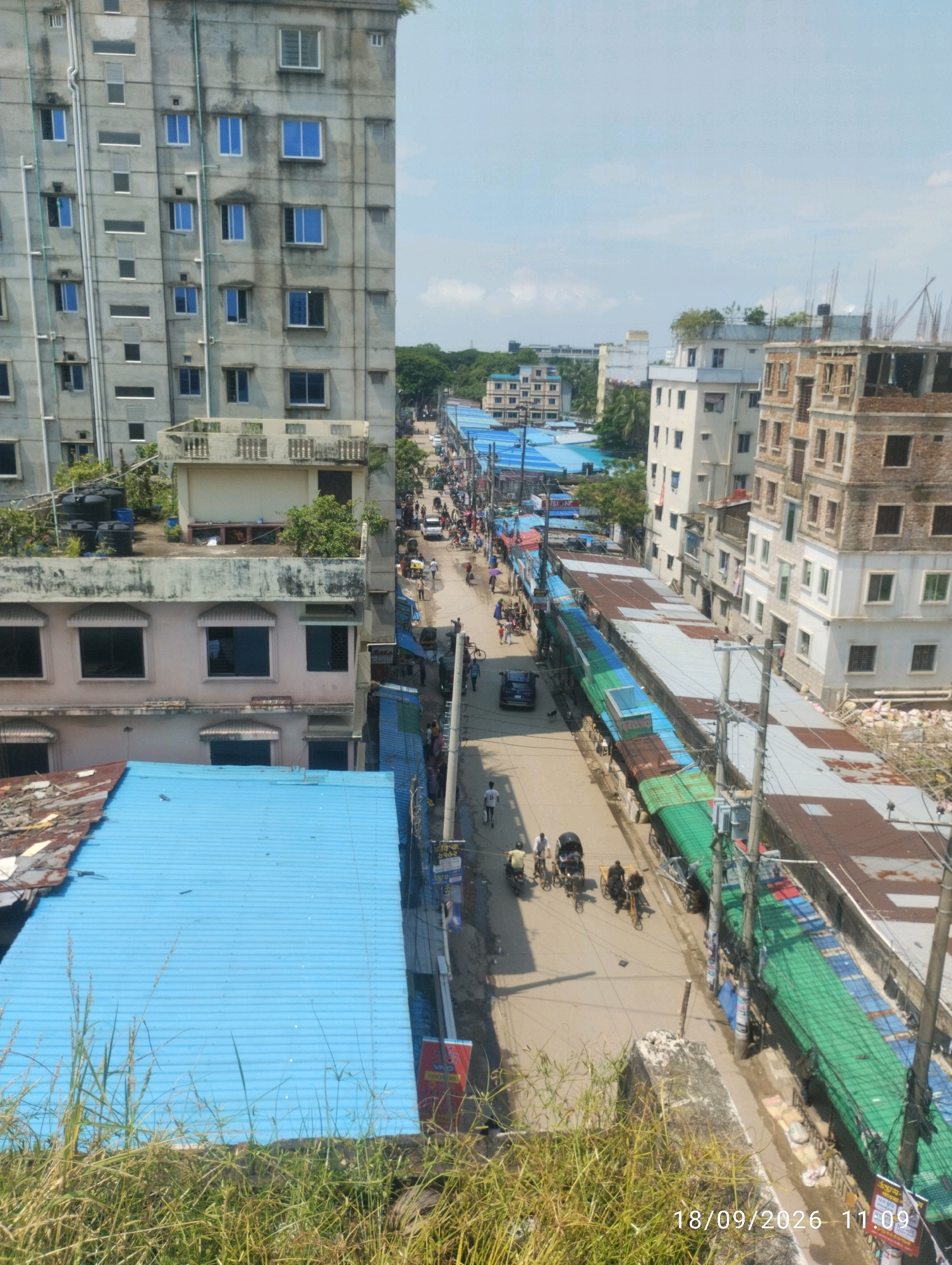 This image depicts a busy street scene in Chattogram, Bangladesh, on September 18, 2026, at 11:09 AM. The perspective is from an elevated position, looking down onto a street flanked by multi-story buildings and a market area.
On the left side of the street, there is a tall, grey, concrete building with numerous windows. Below this building, a lower, light-colored building with arched windows is visible. The foreground on the left features a large, bright blue corrugated metal roof extending across the lower portion of the frame, with some grass and dry vegetation growing in front of it.
The street itself is paved and appears to be bustling with activity. Several people are walking and riding bicycles and rickshaws. There are also a few cars and motorcycles. Along the right side of the street, a long row of market stalls or shops with corrugated metal roofs in various colors, including green, blue, and rust, are set up. Some of these structures appear to be under construction or in disrepair. Behind these stalls, more multi-story buildings are visible, some appearing modern and others older and under construction. Electrical wires and poles are prominent features along the street. The sky is a pale blue with some scattered clouds.