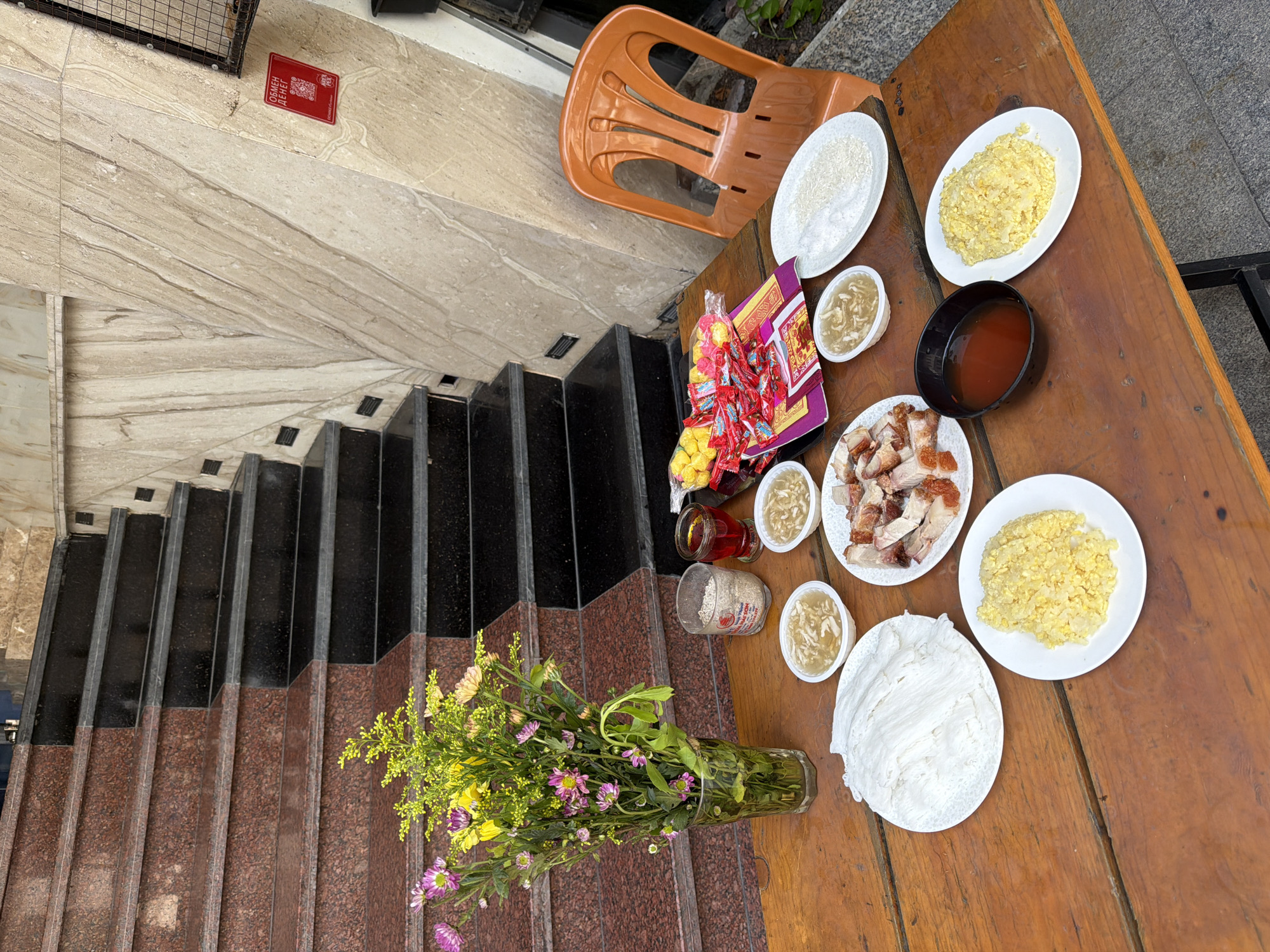 An outdoor table laden with food is set in front of a granite-clad staircase. The table holds several white plates, some filled with rice, and bowls containing a light-colored liquid. In the center, a plate displays sliced roasted meat. A glass vase filled with a colorful bouquet of flowers stands to the left. To the right, an orange plastic chair is positioned next to a wall with a red sign featuring Cyrillic text. The scene appears to be an offering or a meal setup. The location is identified as Nha Trang Ward, Vietnam.
