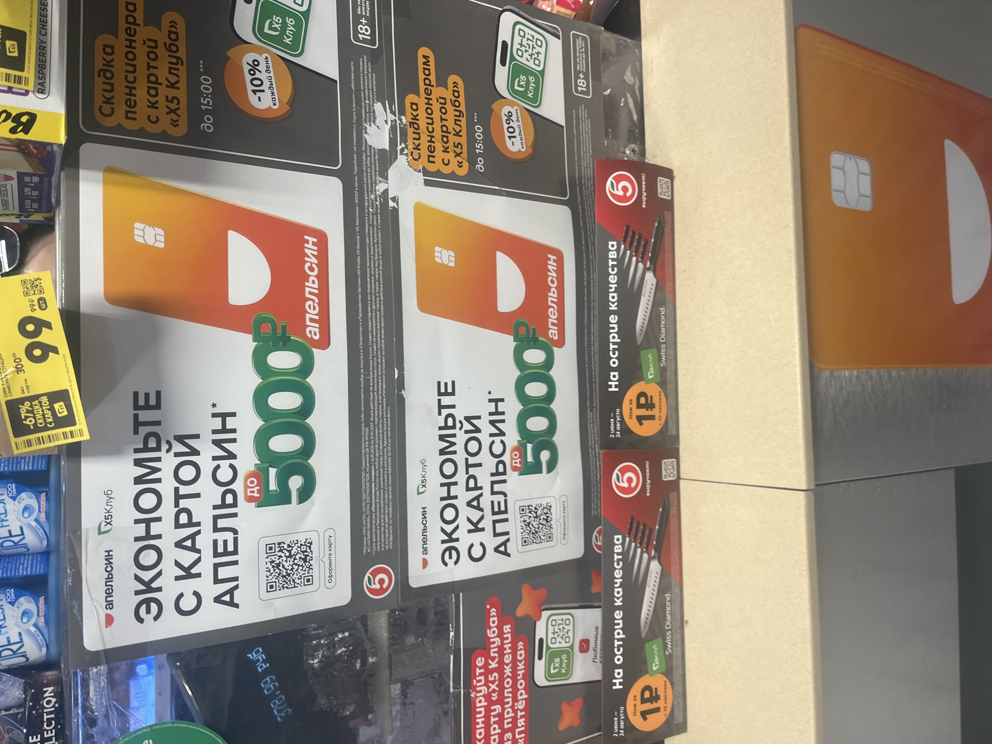 The image is a close-up, eye-level shot of a retail display in Moscow, Russia. The display promotes savings with an "X5 Club" card from "Apelsin." A large graphic features an orange and yellow credit card with a smiling face design. Text on the display highlights "Save with the Apelsin card" and offers up to 5000 rubles in savings. Another section advertises a "Pensioner discount with the X5 Club card" offering 10% off daily until 15:00. Below, a promotion for "Sharp quality" features a set of knives for 1 ruble with 70 stamps. The background shows shelves stocked with various products, including brightly colored packaging for toiletries or food items. The lighting appears to be artificial store lighting. There are no people visible in the image. The overall scene is a point-of-sale display designed to attract shoppers with discount offers.