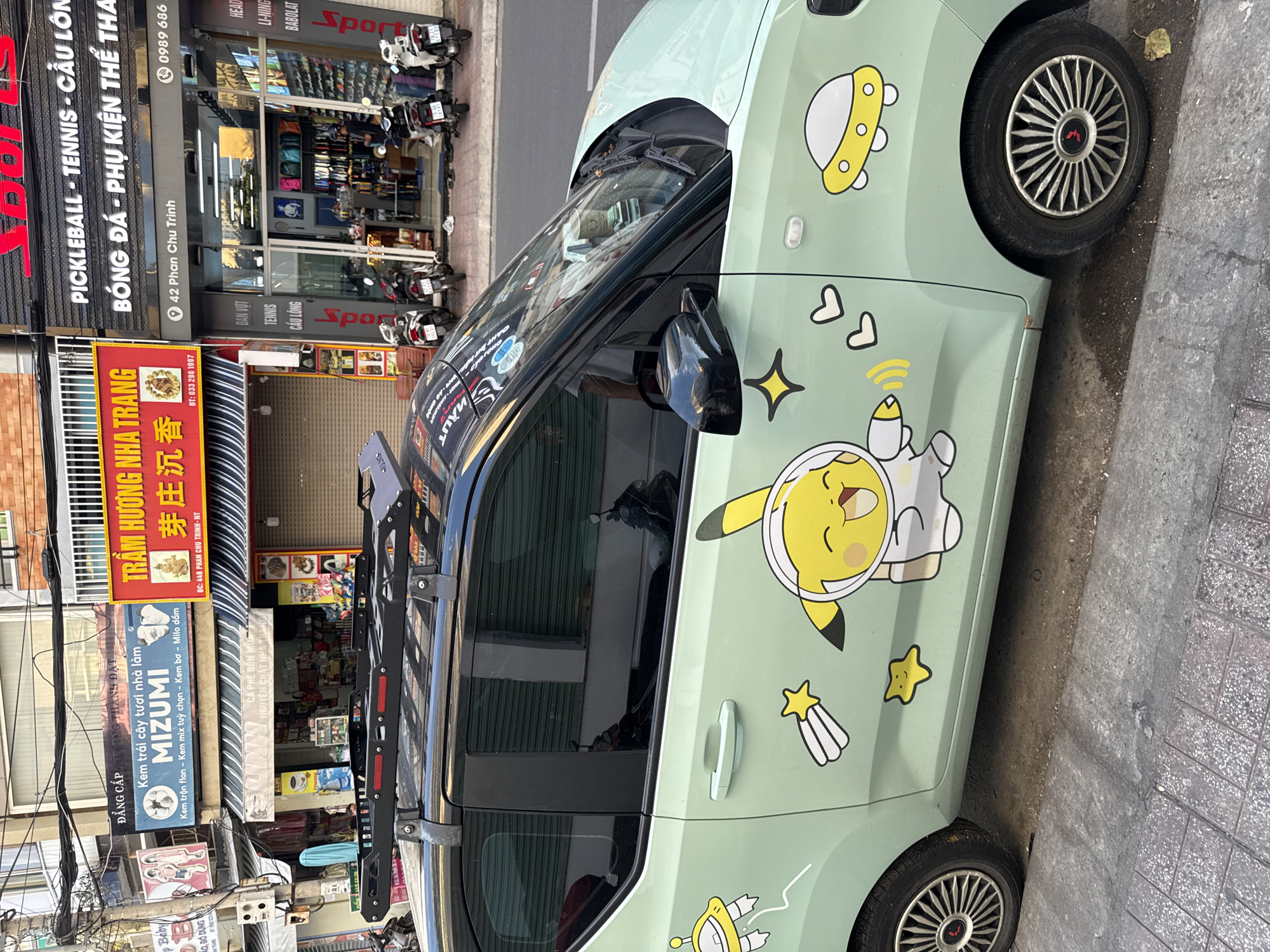 The image shows a pale green car adorned with cartoon stickers of Pikachu in a spacesuit, a UFO, and stars. The car has a black roof rack and tinted windows. It is parked on a city street in Nha Trang, Vietnam, with several shops and buildings visible in the background. One shop, labeled "TRÁM HƯƠNG NHA TRANG," appears to be a local business. Other shops include a sports store labeled "SPORTS" which sells pickleball, tennis, and badminton equipment, and another shop selling Mizumi products. Several motorcycles are parked on the sidewalk in front of the shops. The street is paved with concrete and cobblestones.