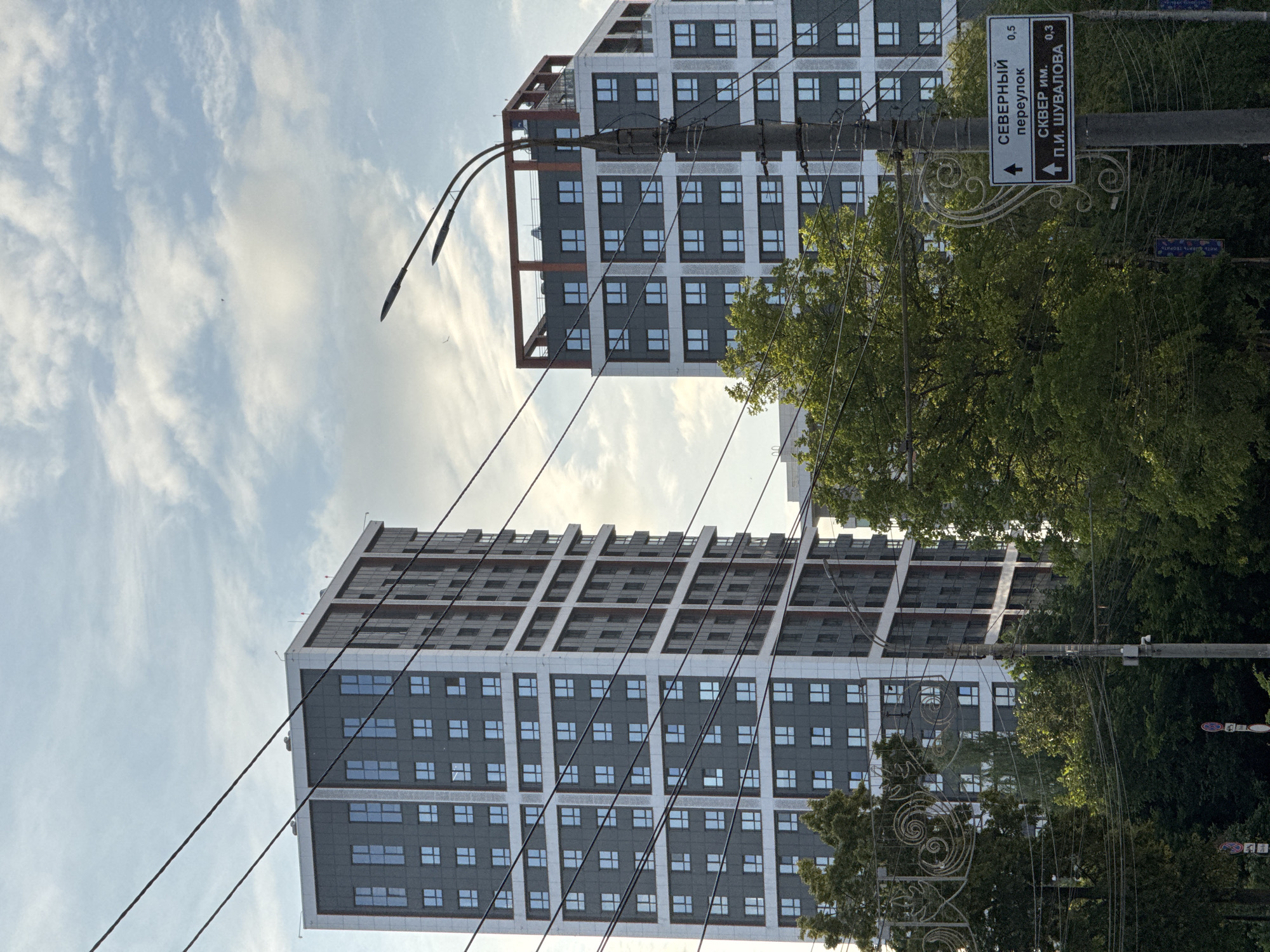In Izhevsk, Russia, two modern apartment buildings stand tall against a cloudy sky. The building on the left is taller and narrower, while the one on the right is shorter and wider, with a distinctive red frame at the top. A utility pole with a signpost is visible in the foreground, indicating "Severny pereulok" (Northern alley) and "SKVER im. P.I. Shuvalova" (P.I. Shuvalov's square) with distances. Lush green trees partially obscure the view of the buildings. Overhead power lines crisscross the sky, adding to the urban landscape. The scene appears to be captured during the daytime, with soft natural light illuminating the surroundings.