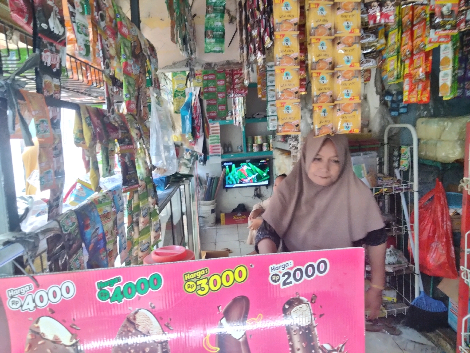 The image depicts a street-side stall in Suka Sari, Indonesia, selling assorted snacks and ice cream. A woman wearing a light brown hijab and a dark top is visible, positioned behind a pink advertisement displaying ice cream bars and prices in Indonesian Rupiah: 4000, 3000, and 2000. The stall is densely packed with various packaged goods hanging from the ceiling and shelves. Behind the woman, a television screen shows a colorful image. To the right, a white rack holds more items, and a red plastic bag and blue broom are also visible. The overall environment suggests a bustling, small retail setting.
