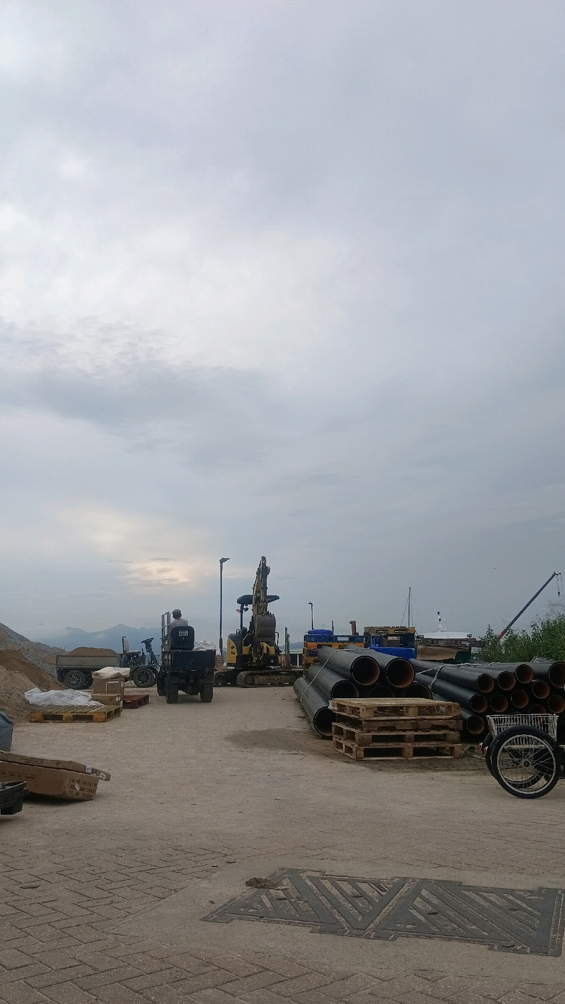 The image depicts an outdoor industrial area under an overcast sky. In the foreground, a paved area with brick-like patterns transitions to a concrete surface. Several large, black pipes are stacked on wooden pallets to the right. A bicycle with a basket is parked nearby. Further back, a yellow excavator is positioned. To the left of the excavator, a person sits on a dark utility vehicle that is loaded with cargo. Another utility vehicle with a trailer filled with sand is also visible. A pile of sand and gravel is situated at the far left. In the background, there are some buildings, a crane, and boats on the water, suggesting a harbor or waterfront location. The overall impression is a construction or storage site. The image does not provide any specific clues to indicate it is located in Hong Kong, China.