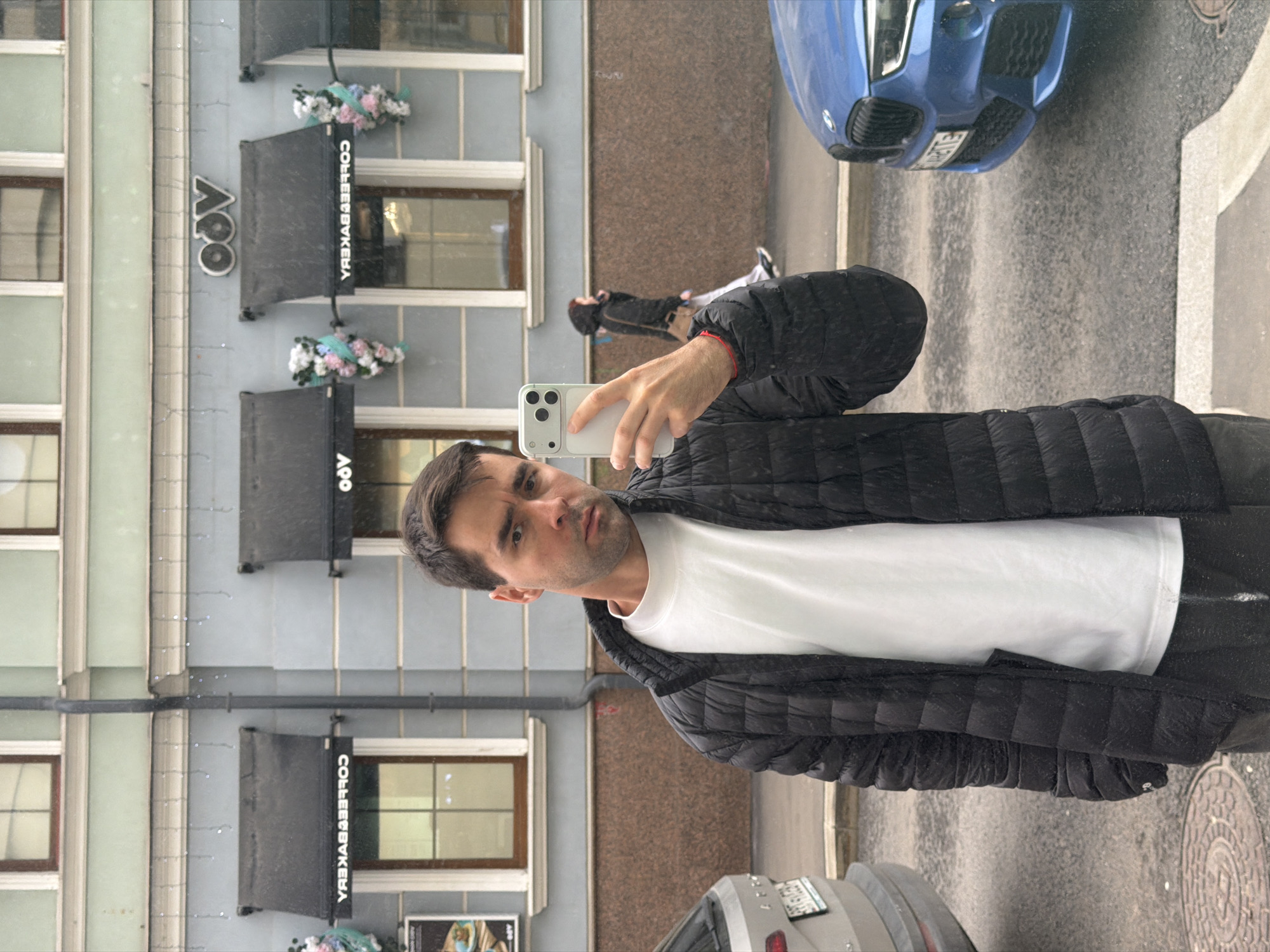 A man is captured in a selfie on an urban street in Moscow, Russia. He is positioned in the foreground, holding a light-colored smartphone with his right hand, looking directly into the camera with a neutral expression. He has dark hair and is wearing a white t-shirt underneath a black puffer jacket, indicating cool weather.
The background features a light blue-grey building on the left, with multiple windows and architectural details. Several dark awnings extend from the building, each bearing the reversed white text "COFFEE&BAKERY." Above one awning, a logo or name "Ooo" is visible. Decorative floral arrangements in pink, white, and blue hues hang beneath the awnings.
To the man's right, a paved street or sidewalk leads into the background. A blue BMW car is partially visible in the mid-ground on the street, identifiable by its front grille and part of its license plate, which shows the numbers "185" and the letters "TE", suggesting a Russian registration. Further down the street, another person can be seen walking. A small section of a grey car is also visible in the lower left foreground. The ground appears slightly damp or reflective, indicating a possibly wet or overcast day. The scene is illuminated by natural daylight.