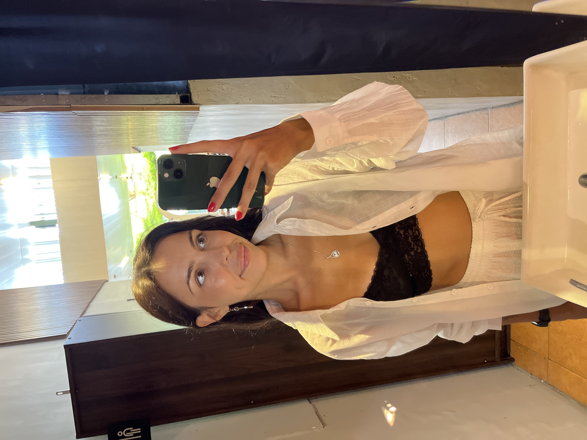 A young woman is taking a selfie in what appears to be a bathroom. She is holding a dark green iPhone in her right hand. The walls of the bathroom are a mix of light gray and dark brown, with a corrugated metal-like material present. A white sink is visible on the right side of the frame.
The woman is wearing a white button-down shirt over a black lace top. She is wearing a silver necklace and earrings. She is smiling at the camera.
Without additional information, I cannot confirm if the location is Kuta Selatan, Indonesia.