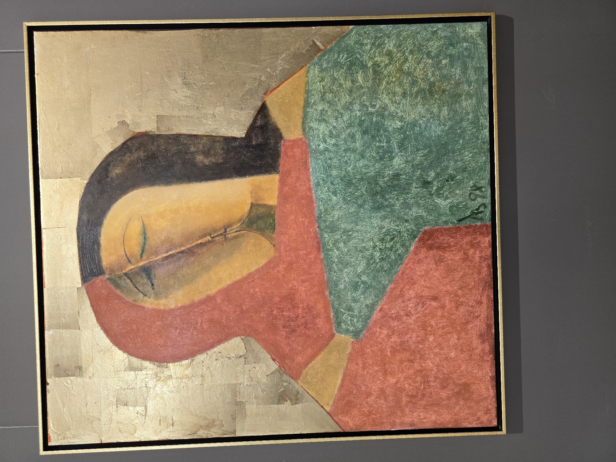 A close-up shot captures a framed painting with a gold leaf textured background. The artwork itself is an abstract depiction of two faces in profile, seemingly embracing or kissing. The left figure is rendered in shades of red and yellow, while the right figure is primarily in yellow and dark brown. They are partially obscured by overlapping blocks of red, green, and gold. The bottom right corner of the painting bears a signature and the year "1998". The painting is displayed on a dark gray wall, suggesting it is in a gallery or exhibition space. The image's metadata indicates it was taken on May 16, 2023, at the National Art Museum of the Republic of Belarus in Minsk.