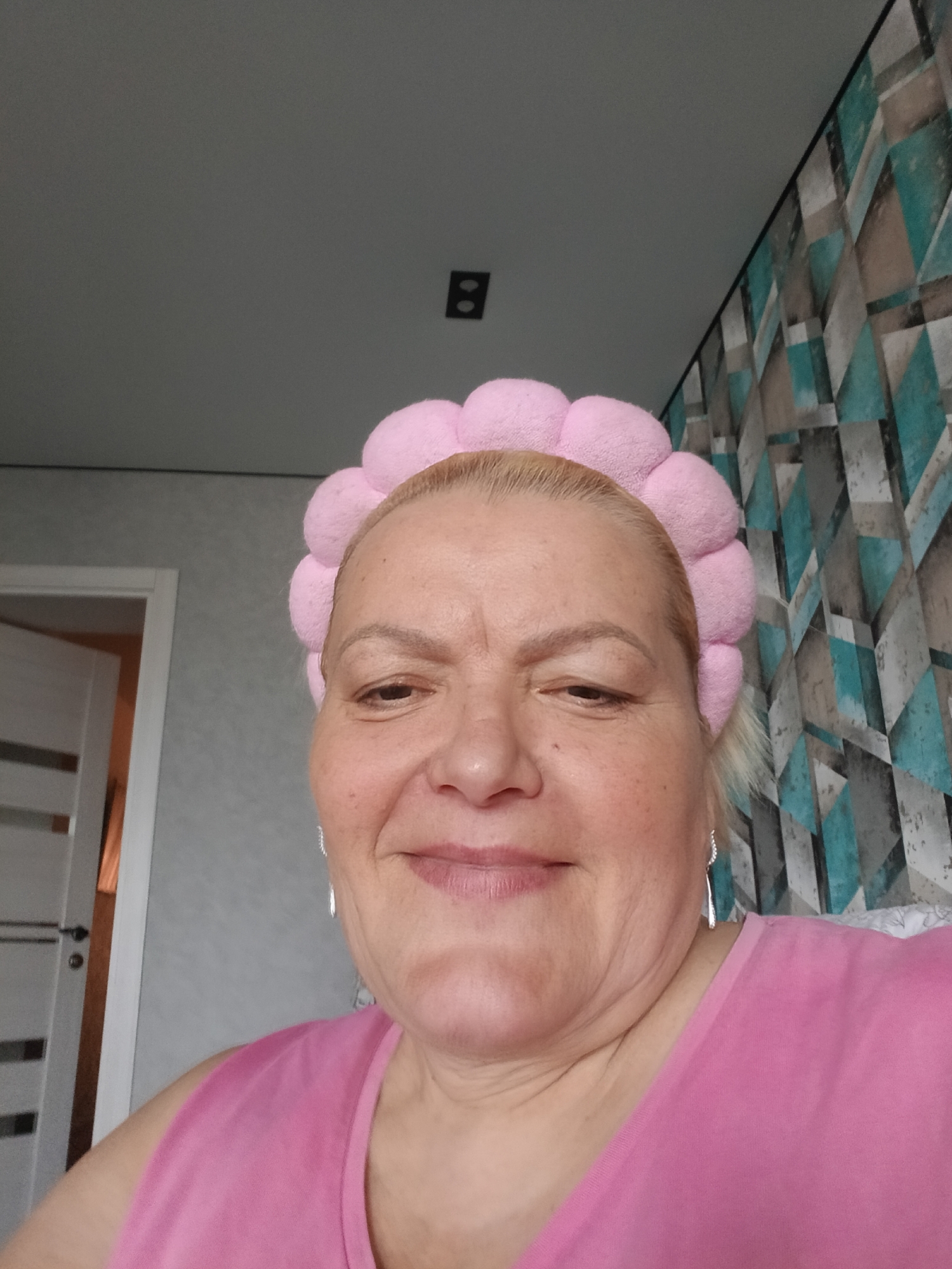 A medium close-up shot of a middle-aged woman with fair skin and blonde hair wearing a pink plush headband and a pink sleeveless shirt. She is smiling slightly. She has silver teardrop earrings. The background includes a wall with a geometric pattern of blue, gray, and white, a light gray wall, a door frame with a partly open white door, and a black electrical outlet. The location is Murom, Russia.