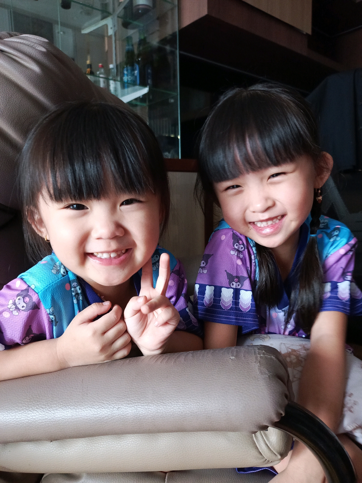 Two young girls, appearing to be of Asian descent, are positioned side-by-side in the foreground of the image. Both are smiling and looking towards the camera. The girl on the left has her hands clasped in front of her, with her right hand extended to form a peace sign. She is wearing a purple and blue garment with a pattern of cartoon characters. The girl on the right also has her hands clasped in front of her and is wearing a similar garment. Her hair is styled in a braid. They are seated on a beige, possibly leather, upholstered chair or sofa. In the background, a glass-fronted cabinet is visible, containing various bottles and objects. The overall setting suggests an indoor environment, possibly a home, in Curug Sangereng, Indonesia.