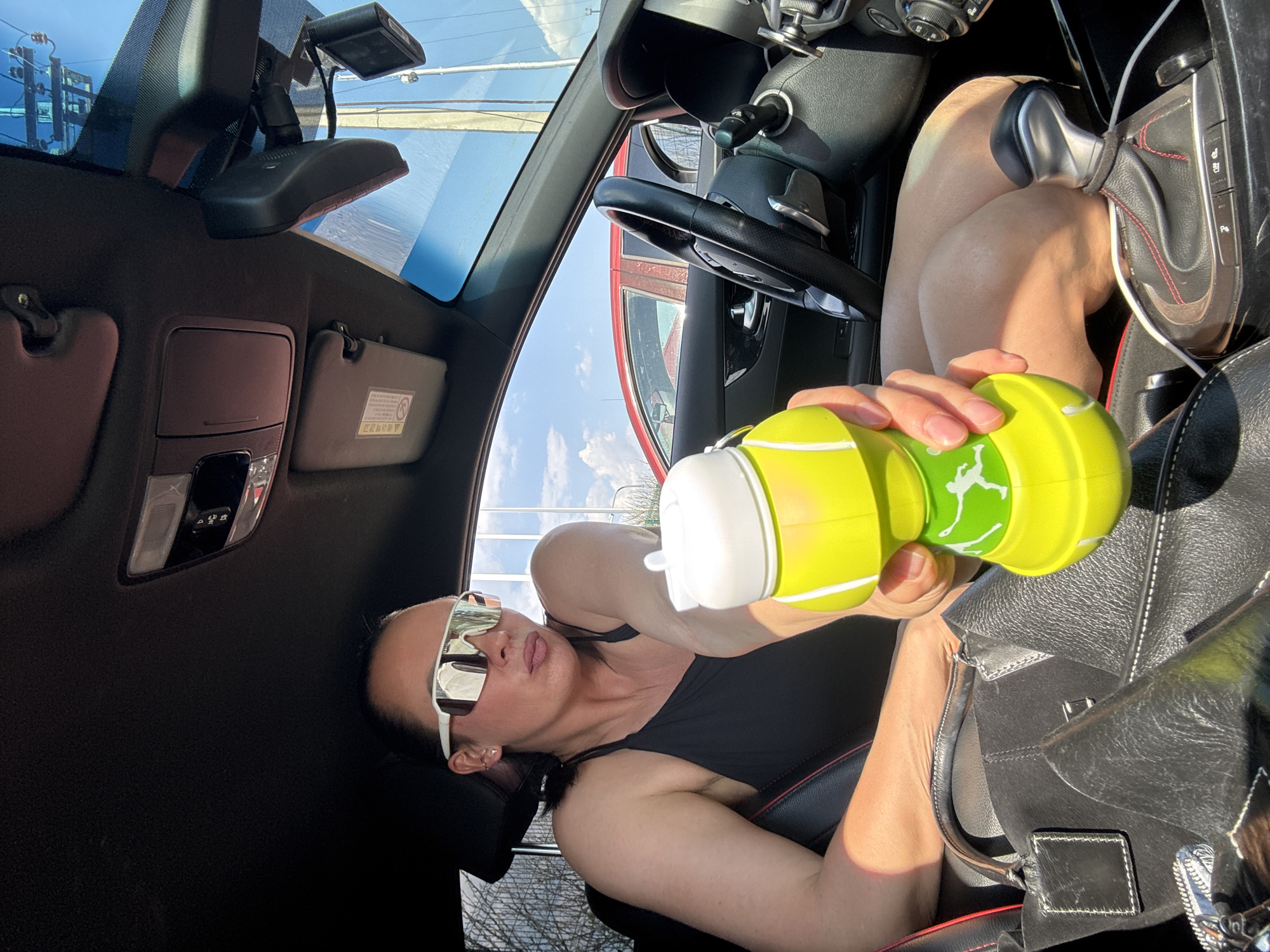 A woman is sitting in the driver's seat of a car. She is holding a yellow water bottle with a white cap. She is wearing sunglasses and a black top. A black leather purse is next to her. The car is parked with a view of a partly cloudy sky visible through the windshield. The location is indicated as Прикамский, Russia.