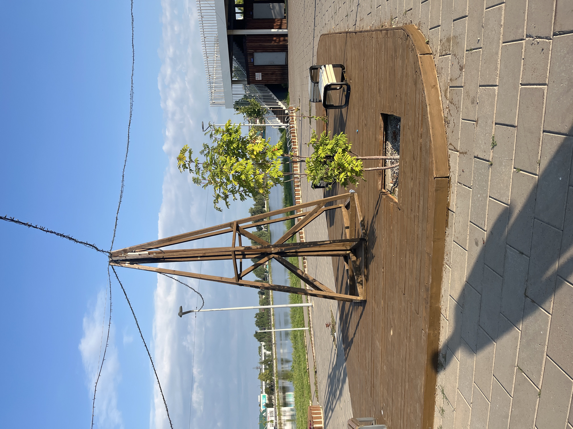The image depicts an outdoor scene in the city of Novye Turbasly, Russia, likely a park or public area near a body of water. In the foreground, a circular wooden platform features a small tree planted in a square opening. A tall, pyramid-shaped wooden structure, possibly a decorative element or part of a larger installation, stands on the platform. Two benches with black metal frames and light-colored seats are also present on the platform. The platform itself is surrounded by a paved walkway made of rectangular gray stones.
Beyond the platform, a calm body of water, likely a lake or river, is visible, with a grassy bank and some trees lining its edge. In the background, a modern building with white railings and stairs is situated. A streetlamp is visible between the wooden structure and the water. The sky is clear blue with scattered white clouds. A long string of lights is draped from the top of the wooden structure and extends across the frame, suggesting the area may be illuminated at night. The shadow of a person, casting a long, distinct shape, falls across the paved walkway in the lower left portion of the image. The overall impression is that of a pleasant, public recreational space.