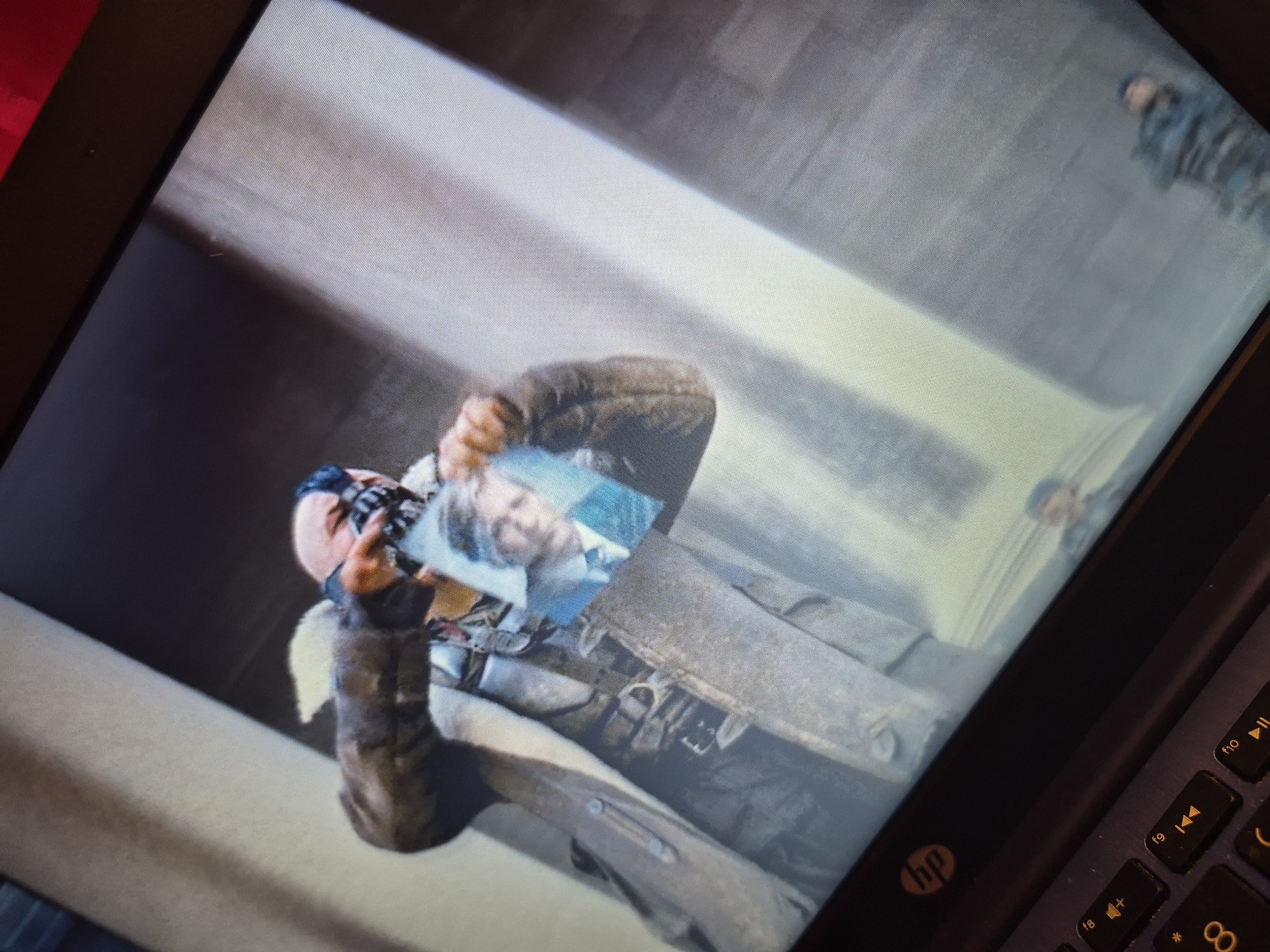 The image shows a close-up, angled view of a laptop screen displaying a scene from the movie "The Dark Knight Rises." A character, identified as Bane due to his mask, is visible in the center of the screen. He is wearing a heavy, dark coat and is holding a torn photograph of a man in a suit and tie. The background on the screen appears to be a large, stone architectural structure, possibly pillars and a building. The lower portion of the image shows the keyboard of the laptop, including several function keys and a visible "hp" logo. The scene depicted on the screen is set in an urban environment, but there is no indication of the specific city of Sandwell, United Kingdom, nor any elements that would allow for its identification.