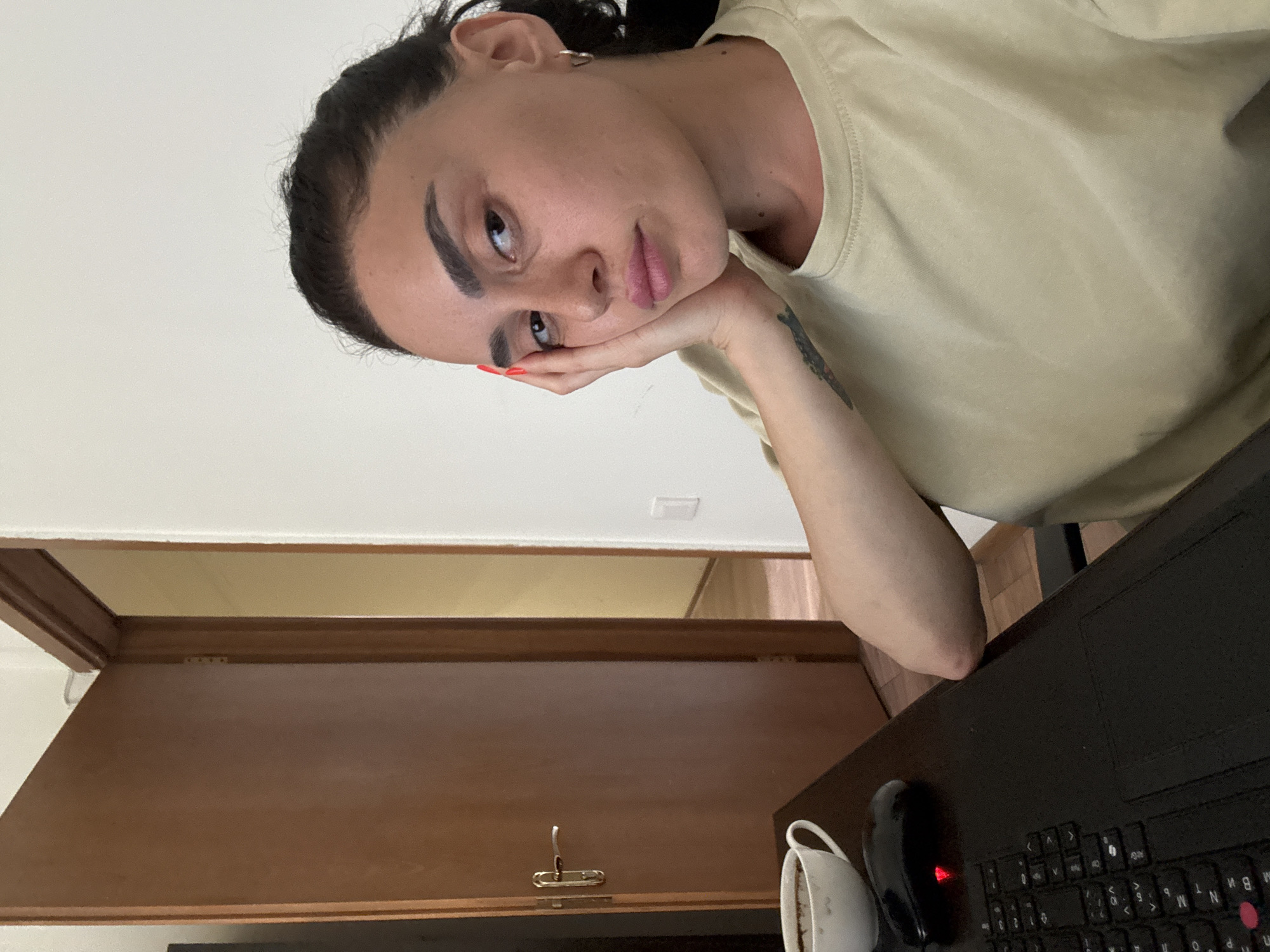 A young woman with dark hair, pulled back in a bun, leans her head on her hand. She has dark eyebrows and is wearing a tan t-shirt. She is sitting at a desk with a laptop and a coffee cup. A door is visible behind her, slightly ajar, revealing another room. The overall setting suggests an indoor environment, possibly a home or office, and the woman appears to be tired or bored. There is no indication of the location being Tbilisi, Georgia.