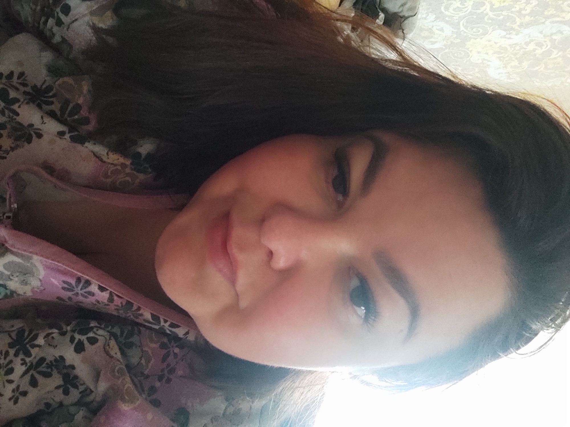 This image depicts a single individual, an adult female, in a close-up, head-and-shoulders portrait, taken indoors within the city of Chaikovsky, Russia. The subject has dark, medium-length hair, which is parted on the left and partially obscures the left side of her face. Her complexion is fair, with a visible flush on her cheeks. She features defined eyebrows and appears to be wearing eye makeup, including extended eyelashes on her visible right eye. Her right eye is open and directed towards the camera, while her left eye is partially closed, creating a winking or squinting expression. Her lips are closed, forming a subtle, upward curve. She is dressed in a garment with a floral pattern, featuring purple, green, and dark floral motifs on a lighter fabric, with a pink trim visible at the neckline. The background on the upper left side shows a light-colored wallpaper with an ornate, possibly damask-style, pattern. To the upper right, a strong, overexposed light source, likely a window, illuminates the scene. Indistinct dark shapes are visible in the upper left background.