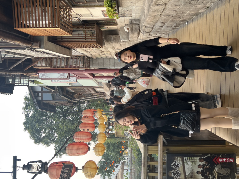 The image captures a bustling pedestrian walkway within Hongyadong Commercial Street, Yuzhong District, Chongqing, China. Traditional multi-story Chinese buildings with wooden structures, latticework windows, and stone foundations line the right side of the wooden-planked path. Overhead, a series of red and yellow paper lanterns hang, alongside a black sign for a "茶舍" (Tea House) on the left.
In the left foreground, a young female with long dark hair, wearing a black hooded sweatshirt, a dark patterned mini-skirt, beige tights, and cream-colored leg warmers, is holding a smartphone, seemingly capturing a photo or video. She carries a black quilted handbag with a gold chain strap. In the right foreground, another young female with long dark hair, dressed in a black long-sleeved top, black wide-leg pants, and white sneakers, walks towards the viewer's right. She has a light pink smartphone mounted on a flexible holder at her chest and carries a black jacket draped over her left arm.
The mid-ground and background feature multiple individuals walking along the path. One person in the mid-ground, wearing an apron, carries a white plastic bag. The scene is illuminated by diffused daylight under an overcast sky, highlighting the architectural details and the flow of pedestrians through this cultural and commercial area.