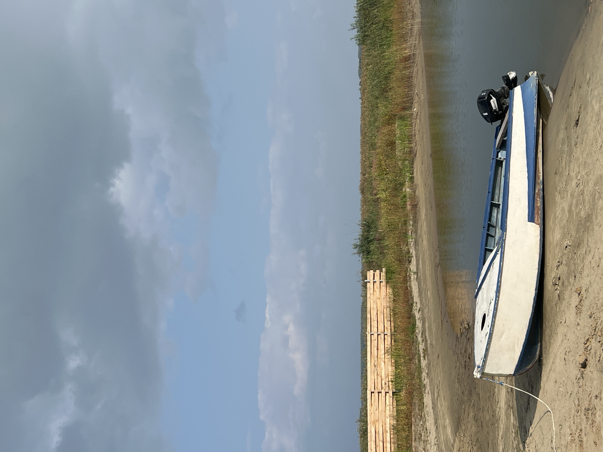 A low-angle, vertical shot captures a serene scene on the shore of the Yenisei River near Novopichugovo, Russia. A small, white and blue motorboat rests partially on the sandy bank, its bow facing left and stern angled towards the water. A black outboard motor is attached to the stern. A white rope trails from the boat's bow onto the sand. Behind the boat, a strip of dry grass and reeds borders the river. Further inland, a stack of freshly cut wooden planks, likely for construction, is neatly piled. The sky above is a mix of pale blue and scattered gray clouds. The overall atmosphere is tranquil, suggesting a peaceful day by the water.