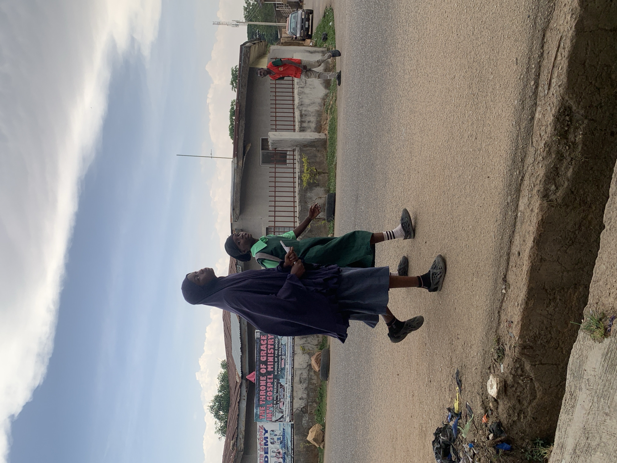 Two girls in school uniforms, one wearing a hijab and navy covering, walk along a paved road. A man in a red shirt walks on the opposite side of the road, near a building with a rusty iron fence. A car is parked further down the street. A religious poster is affixed to a building behind the girls. A concrete curb borders the right side of the road, adjacent to a ditch containing trash. The sky is partially cloudy. The location is Gura Top, Nigeria.