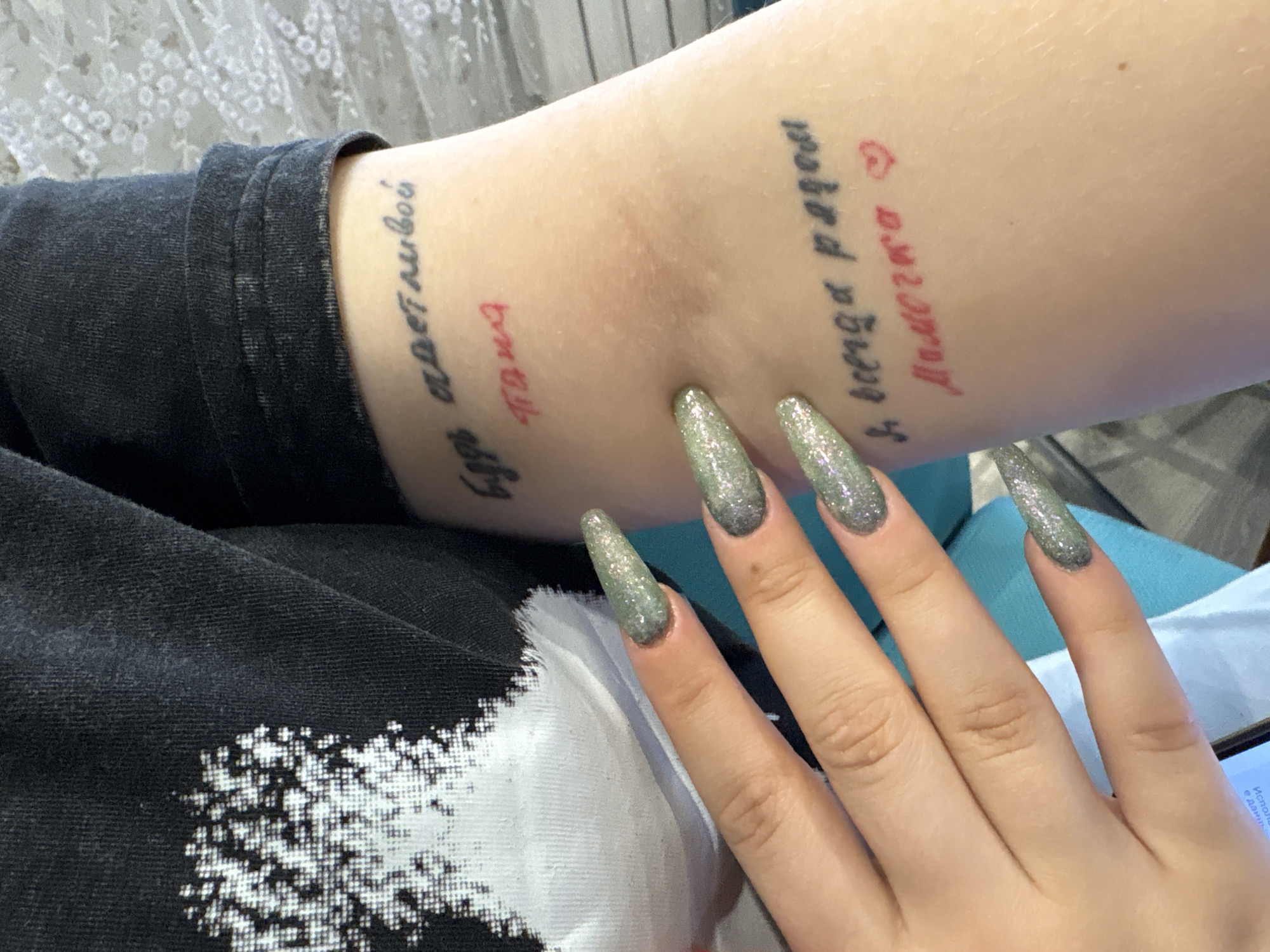 Here is a detailed description of the image:
An arm with several tattoos in Russian is in focus. The tattoos appear to be phrases or words, some in black ink and some in red. The skin of the arm is fair. Part of a black shirt with a white design is visible on the left side of the image. A hand with long, silver glitter nails is resting on the arm. In the bottom right corner, part of a smartphone screen is visible. A teal-colored soft surface is partially visible beneath the arm and hand. The location may be Chaikovsky, Russia, based on the tattoo language, but there is no direct visual information to confirm the city.