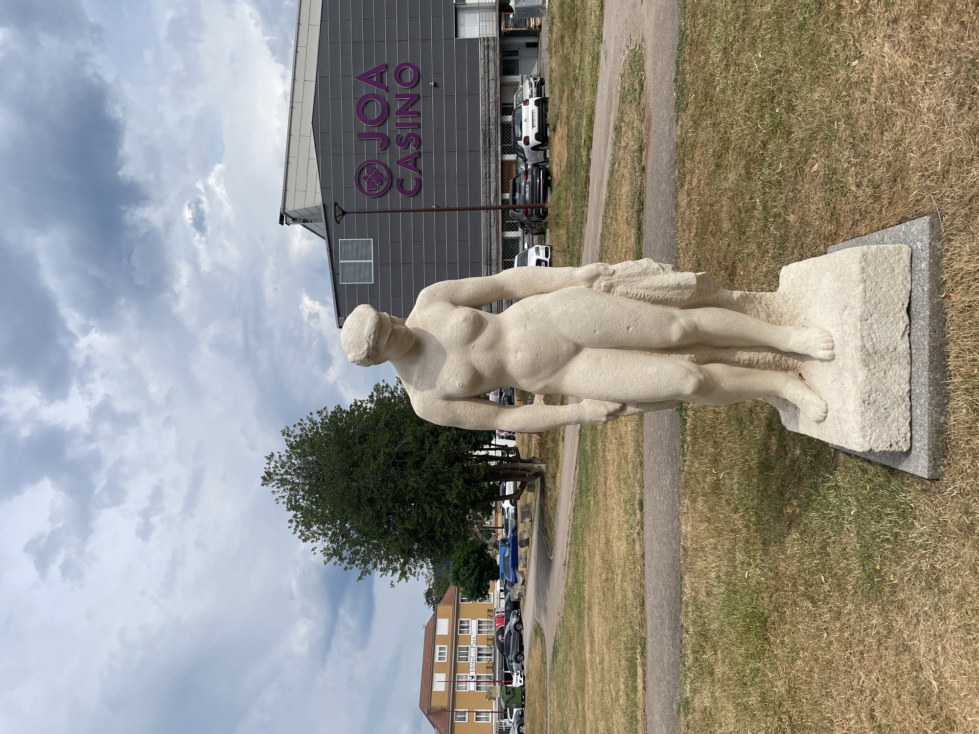 An eye-level medium shot captures a pale stone statue of a nude woman standing in front of a building with the words "JOA CASINO" in large purple lettering. The statue is positioned centrally in the frame, with its head bowed and draped fabric in its right arm. Behind the statue, a paved pathway cuts through a dry, grassy park area. The casino building, a modern, dark grey structure, dominates the background. To the left of the casino, a yellow building with "Hotel Le Paix" signage is visible. Several cars are parked in the lot between the casino and the hotel. The sky above is overcast with scattered clouds, and a large green tree stands to the left of the statue, partially obscuring the hotel. The scene appears to be in an urban outdoor setting, likely a public space in Gérardmer, France, given the prominent "JOA CASINO" signage.