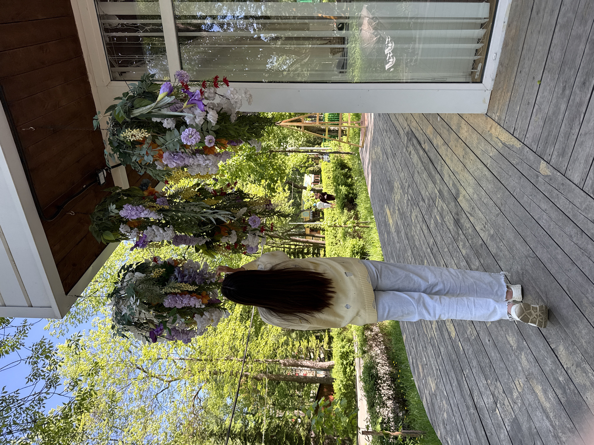 A person with long dark hair is standing on a wooden deck, facing away from the viewer. They are wearing a light yellow sweater with holes and light grey jeans, paired with white and beige shoes. Above them are three hanging floral arrangements, suspended from the roof of what appears to be a building with a large window.
The background features a park-like environment with green trees and glimpses of other structures and people in the distance.
Given the information, the location is most likely Perm, Russia.
