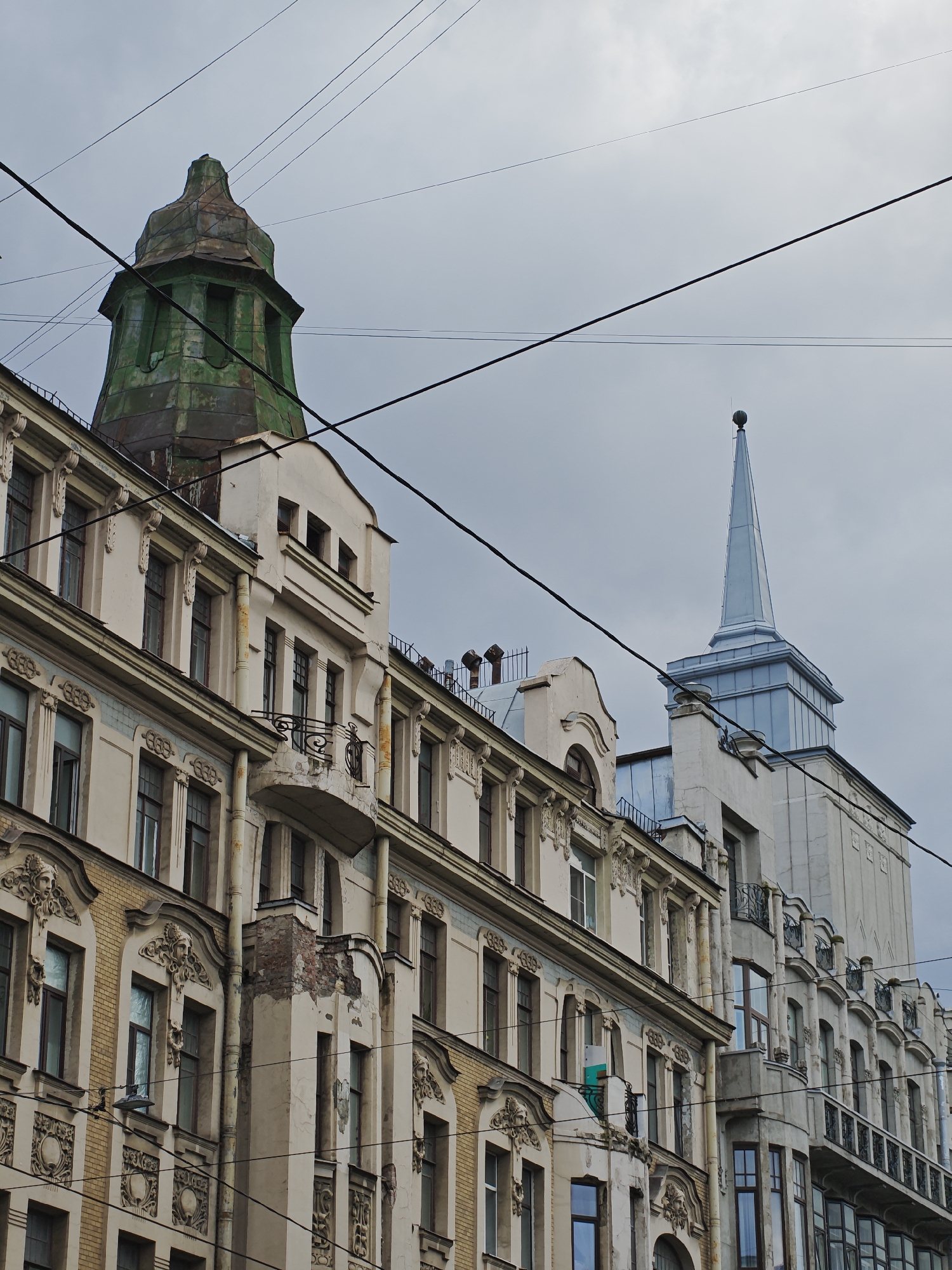This image captures a detail of the architectural facade of buildings in Saint Petersburg, Russia, under a cloudy sky. In the foreground, a cream-colored building with ornate detailing around its windows and balconies dominates the left side of the frame. The facade features decorative elements such as carved faces and geometric patterns. To the left of this building, a portion of a neighboring structure is visible, appearing to be made of yellow brick with cream-colored plaster.
Behind these buildings, and slightly to the right, a taller, lighter-colored building with a distinctive spire rises into the sky. The spire is silver and tapers to a point, topped with a small sphere. To the far left, a distinctive, weathered green tower with a pointed, somewhat conical roof is visible, jutting above the main building. This tower appears to be made of metal and shows signs of age and oxidation.
Multiple dark electrical wires crisscross the sky, obscuring parts of the buildings and the sky itself, suggesting an urban environment. The overall impression is one of historical architecture in a European city, characterized by intricate detailing and a somewhat muted color palette due to the overcast weather. There are no people or animals visible in the image. The scene appears to be a static street view.