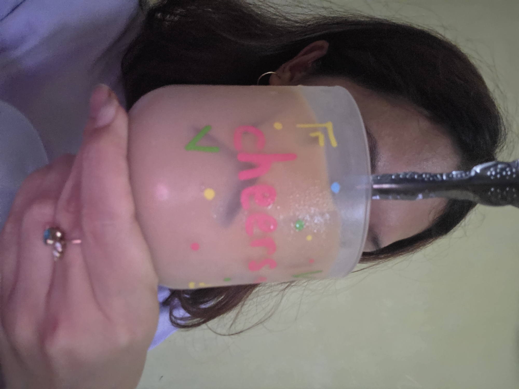 A person is holding a light peach-colored drink with the word "Cheers" written on the cup in pink letters, with green, yellow, and blue confetti dots. The person's hand is holding the cup, and they are wearing a ring with a turquoise stone. A spoon is sticking out of the drink. The person has dark hair and is wearing a light purple shirt. The background is a light yellowish-green. The city is Unknown, Unknown.