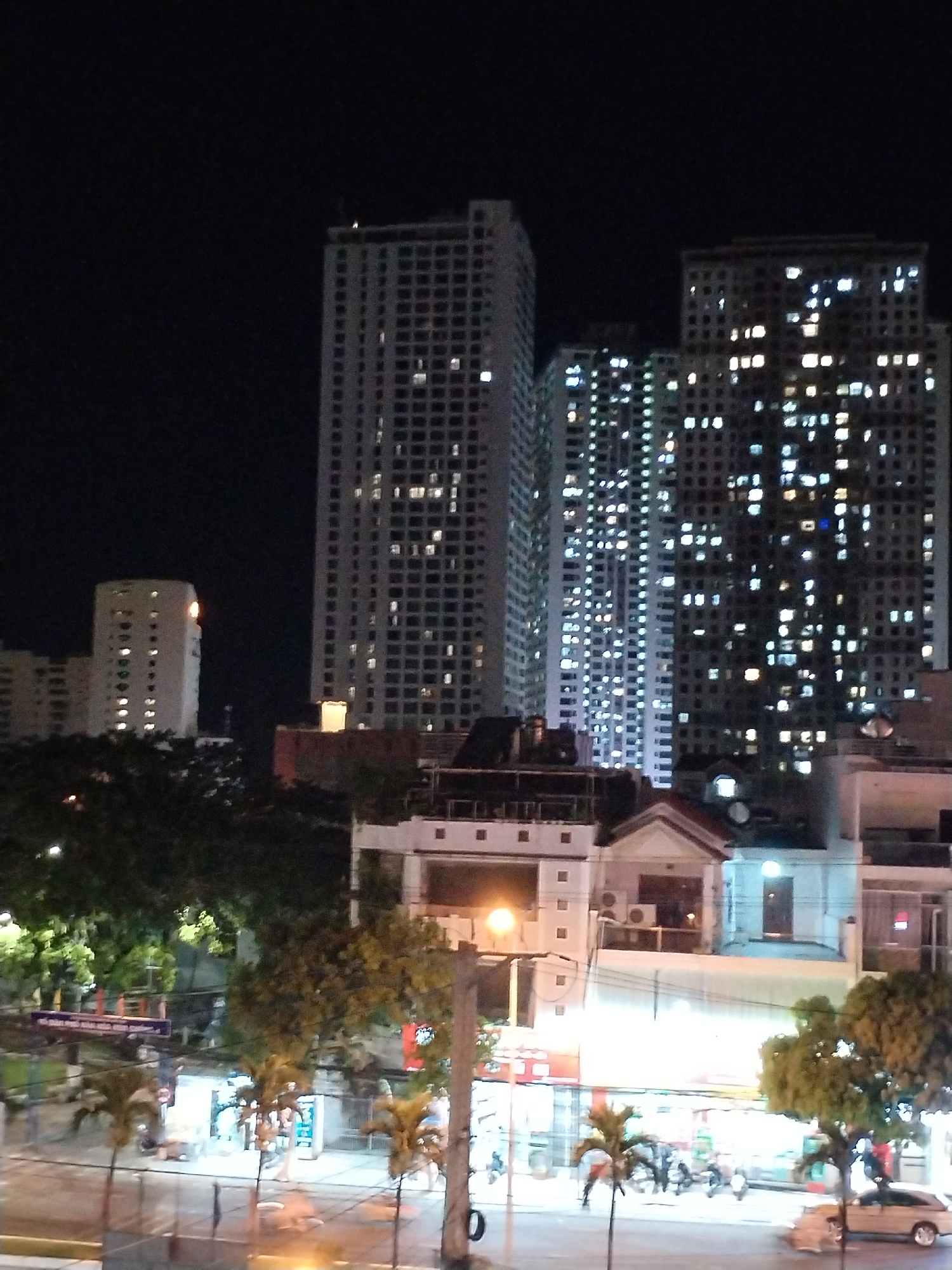 The image shows a nighttime view in Nha Trang Ward, Vietnam. The scene features several tall, illuminated buildings in the background, likely residential or commercial towers, densely packed with windows showing interior lights. The foreground includes lower-rise buildings and street-level activity. There are trees and streetlights, and there's the presence of a small park or plaza. In the street, there are parked vehicles and a few people visible, suggesting a moderate level of activity.