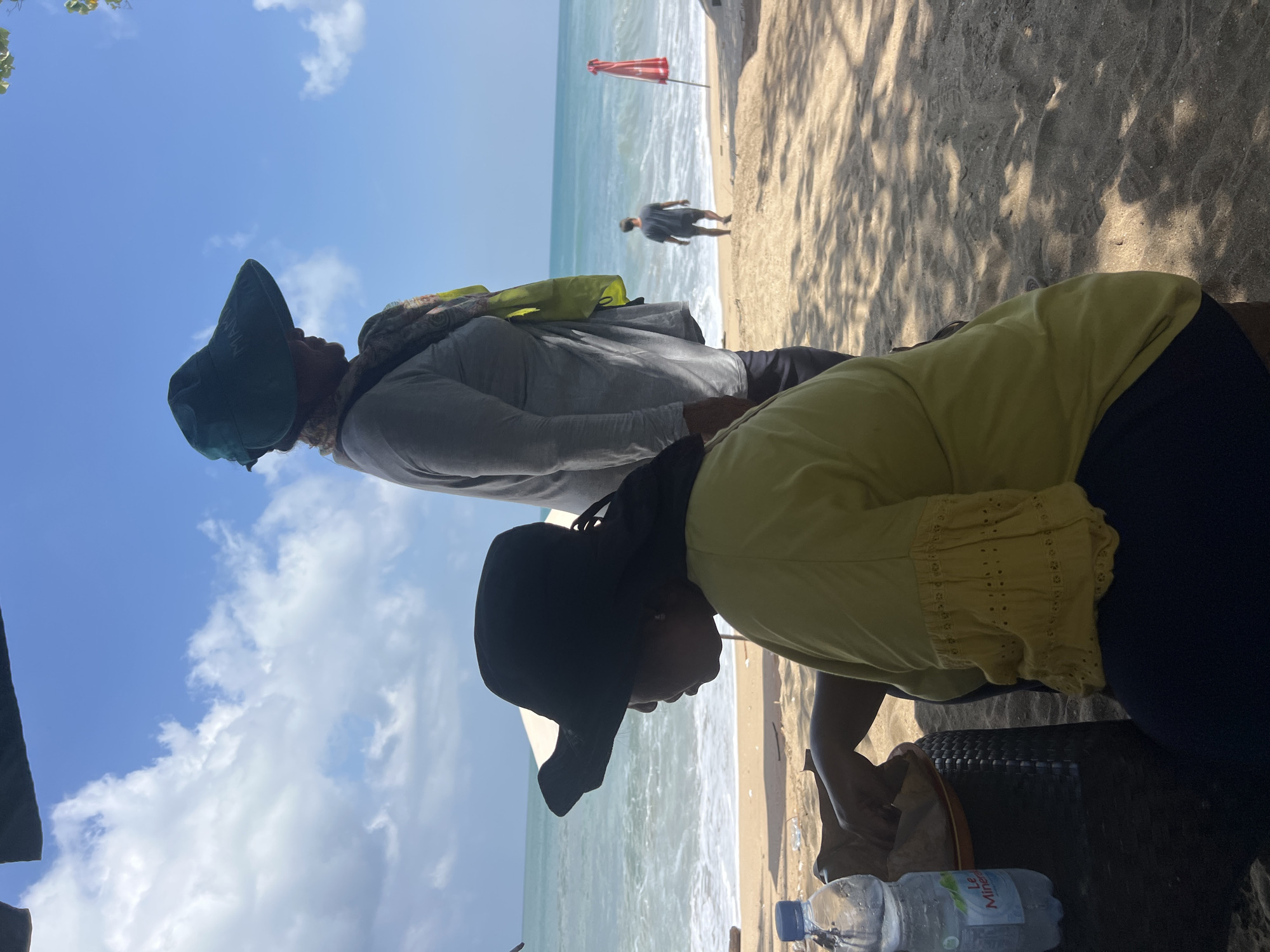 In Kuta, Indonesia, on a sandy beach, two women are seated under a dark canopy. The woman in the foreground is wearing a yellow shirt with ruffled, decorative sleeves and a dark hat. She is sitting next to a black woven table and a bottle of Le Mineral water. To her right, another woman is wearing a gray long-sleeved shirt and a wide-brimmed green hat. She has a colorful scarf draped over her shoulder. Further in the background, a man is walking towards the ocean, and a red umbrella is planted in the sand. The sea is calm with gentle waves. The sky is blue with scattered white clouds. Sunlight casts dappled shadows on the sand.