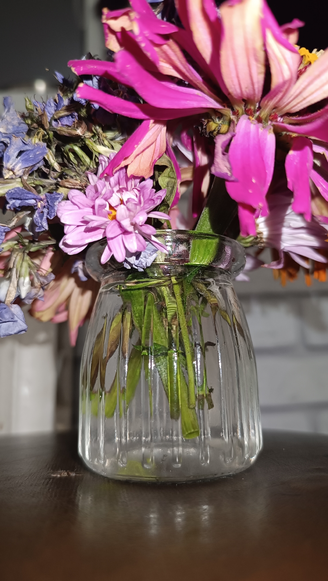 The image shows a close-up of a clear glass vase filled with a bouquet of flowers. The vase has vertical ribbing and is sitting on a dark, reflective surface, possibly a wooden table. The bouquet consists of several types of flowers, including vibrant pink zinnias and delicate purple asters, some of which appear wilted. The stems of the flowers are visible submerged in water inside the vase. The background is slightly out of focus, showing a white brick wall and a blurred white object. There is no discernible indication of the city of Chaikovsky, Russia, in this image. The scene is a still life composition of flowers in a vase.